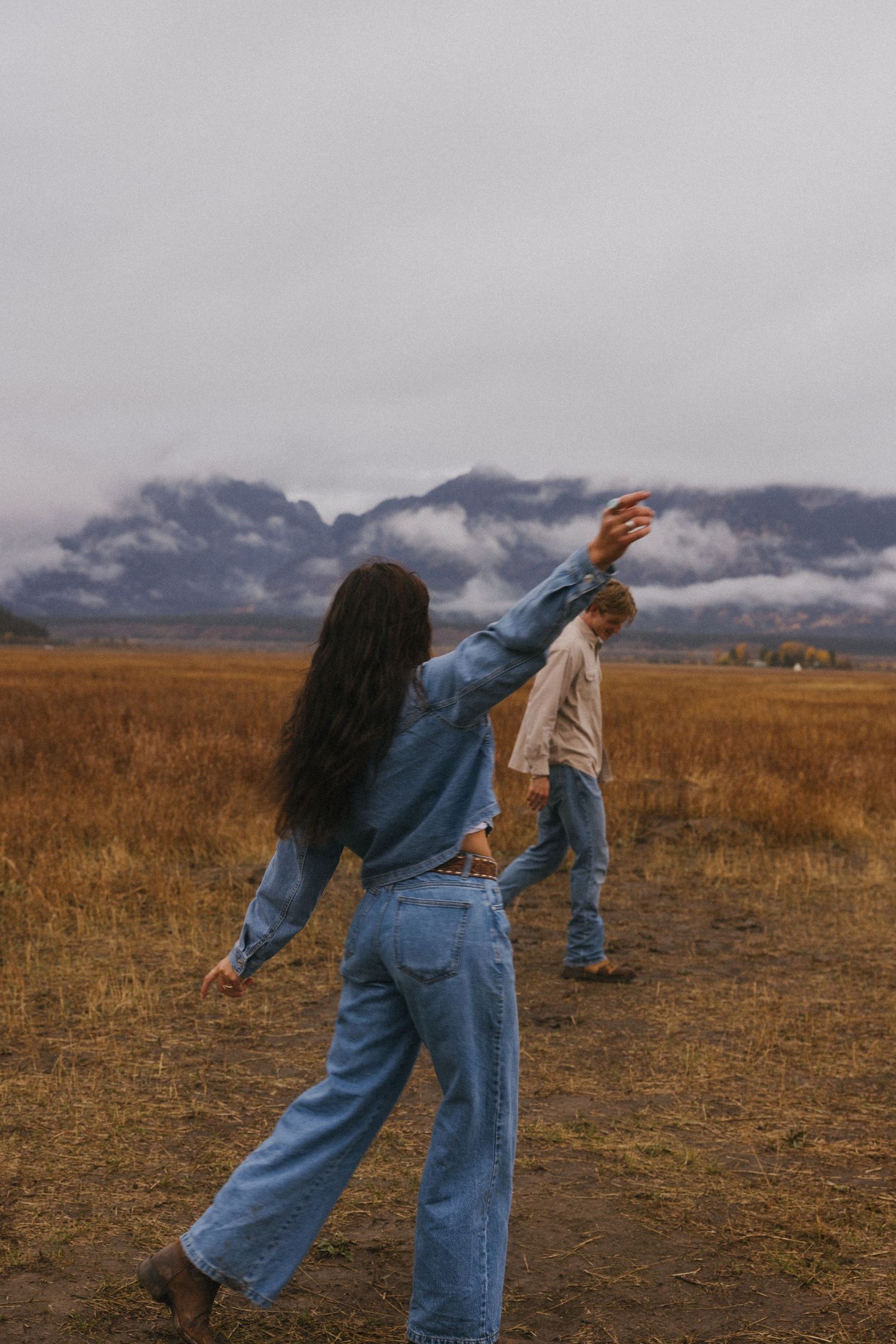 Two people perform expressive dance moves in a field beneath cloudy mountain peaks.