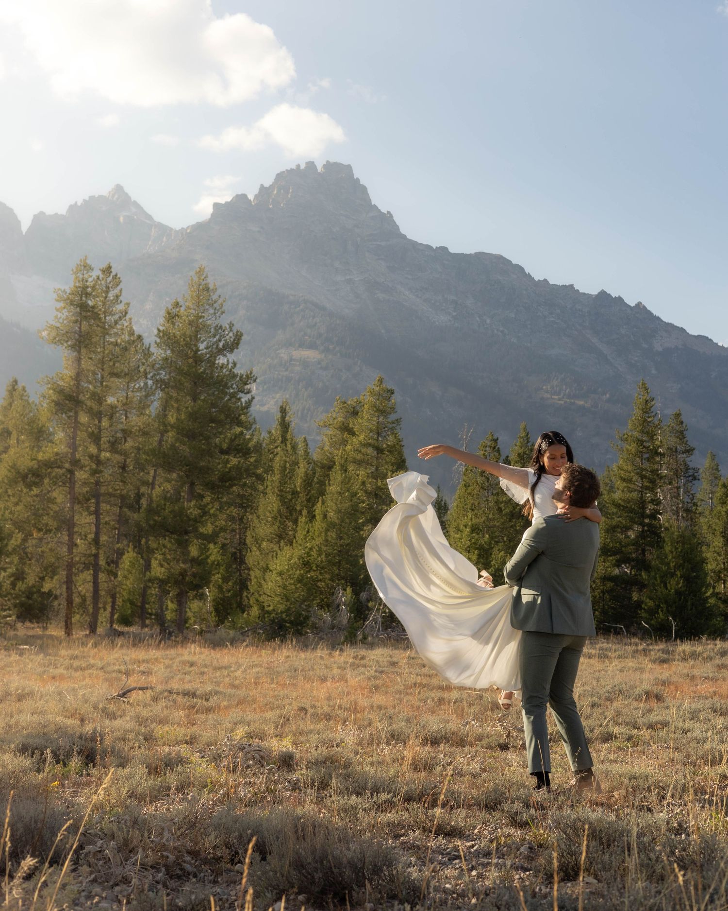 Playful wedding couple dancing in mountain meadow as bride's veil flows in wind.