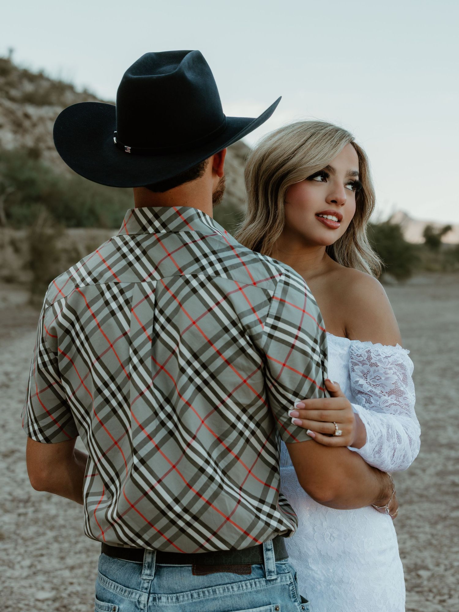 A couple in western attire and white dress pose together against desert landscape.