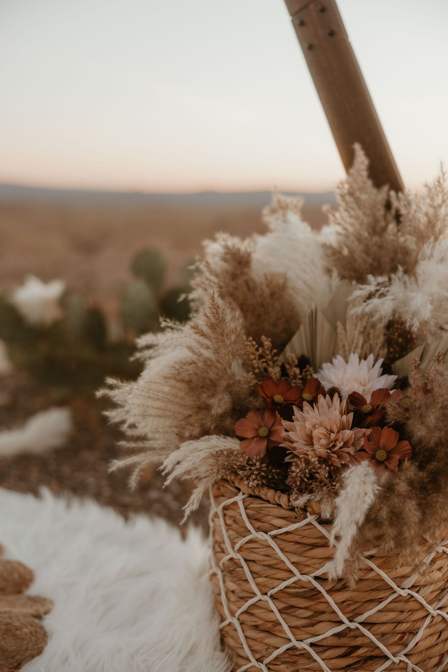 A rustic woven basket filled with dried pampas grass, flowers and autumn-toned floral arrangements against a blurred landscape.