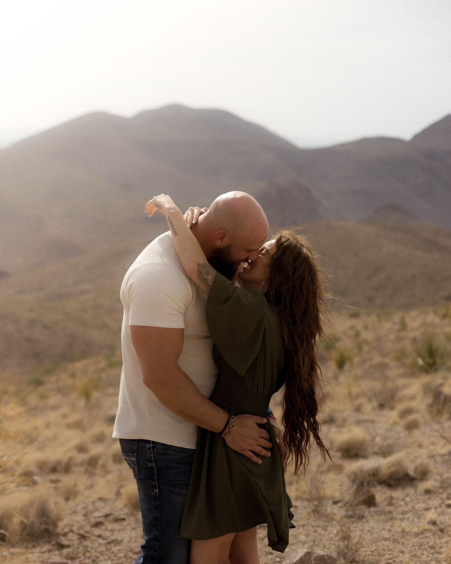 A romantic couple embraces while standing in a desert landscape with mountains in the background.