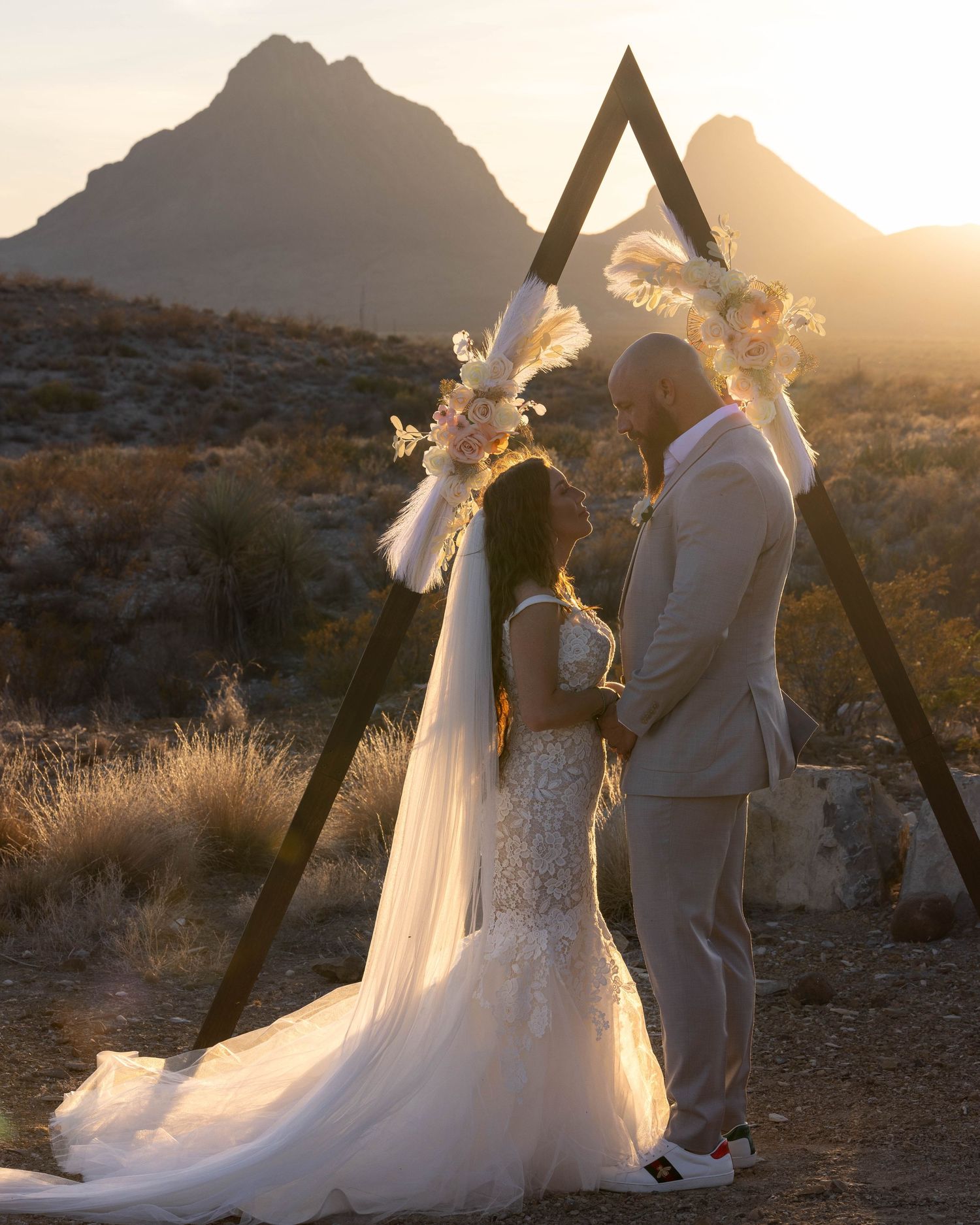 Desert wedding ceremony with wooden triangle arch and flowing white dress against mountain backdrop.