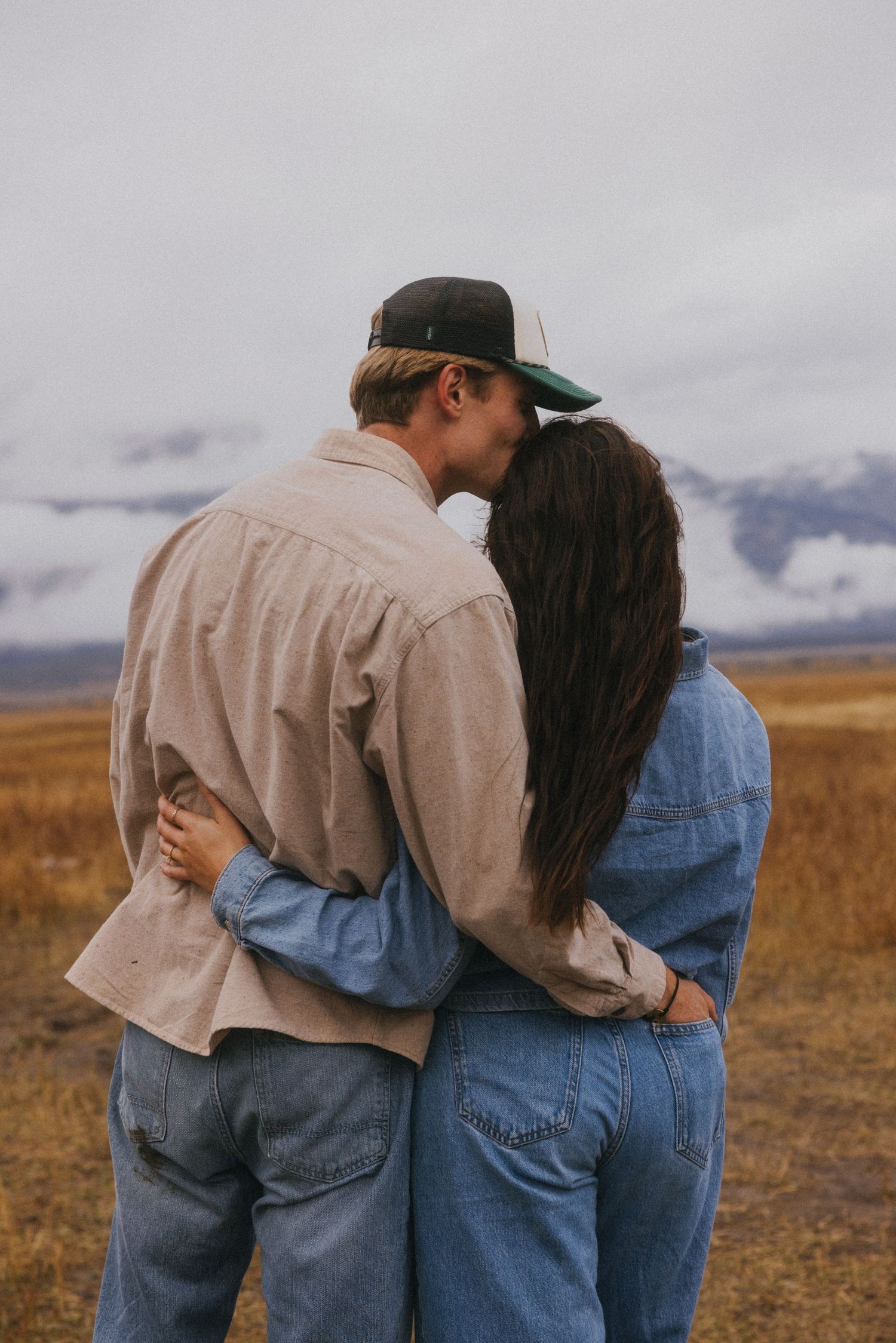 A couple walks together arm in arm across an autumn field with mountains in the distance.