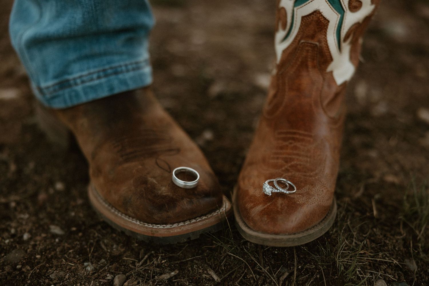Close-up of two pairs of brown cowboy boots with wedding rings on rocky ground.