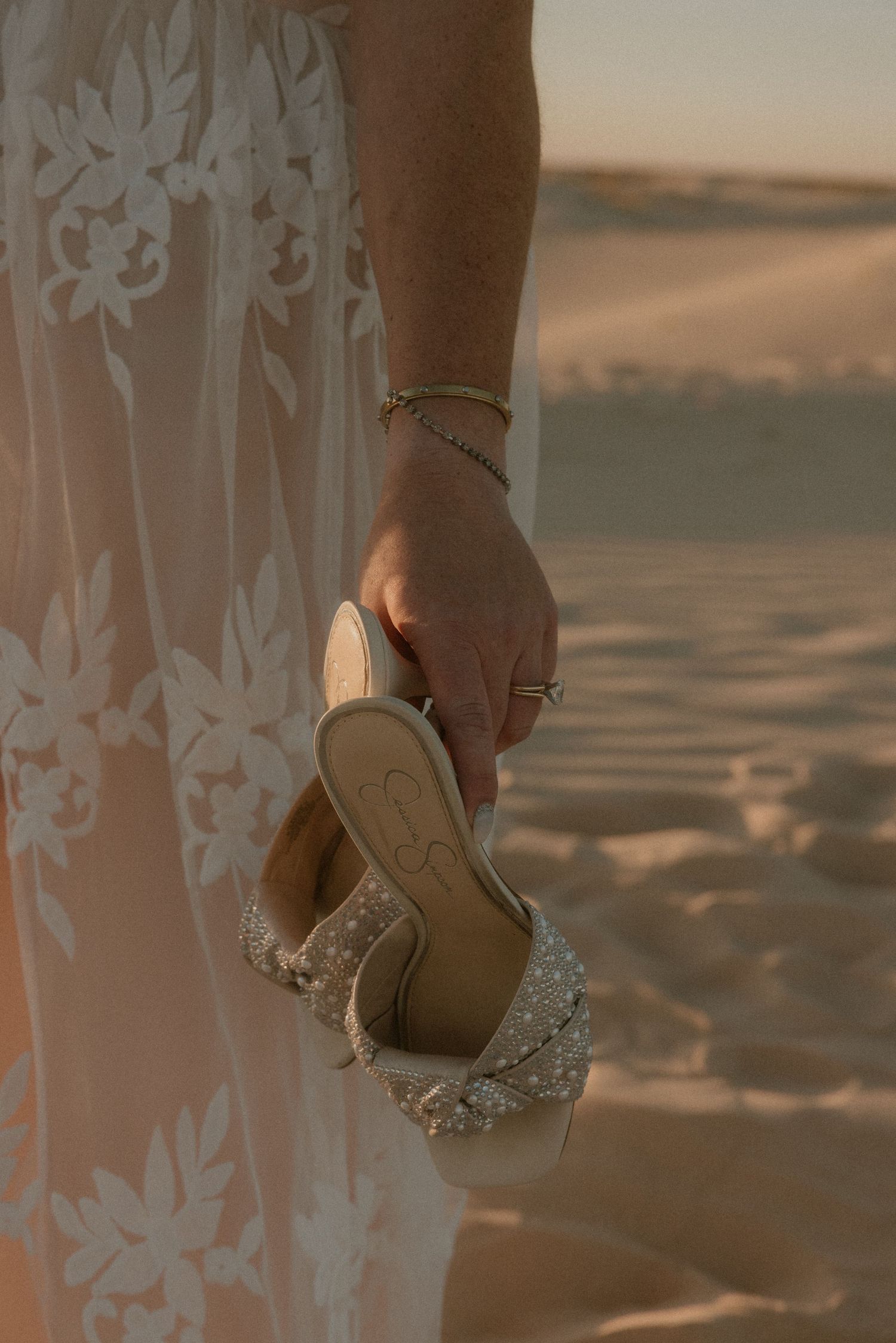 A close-up of a bride holding embellished wedding shoes against desert sand at sunset.
