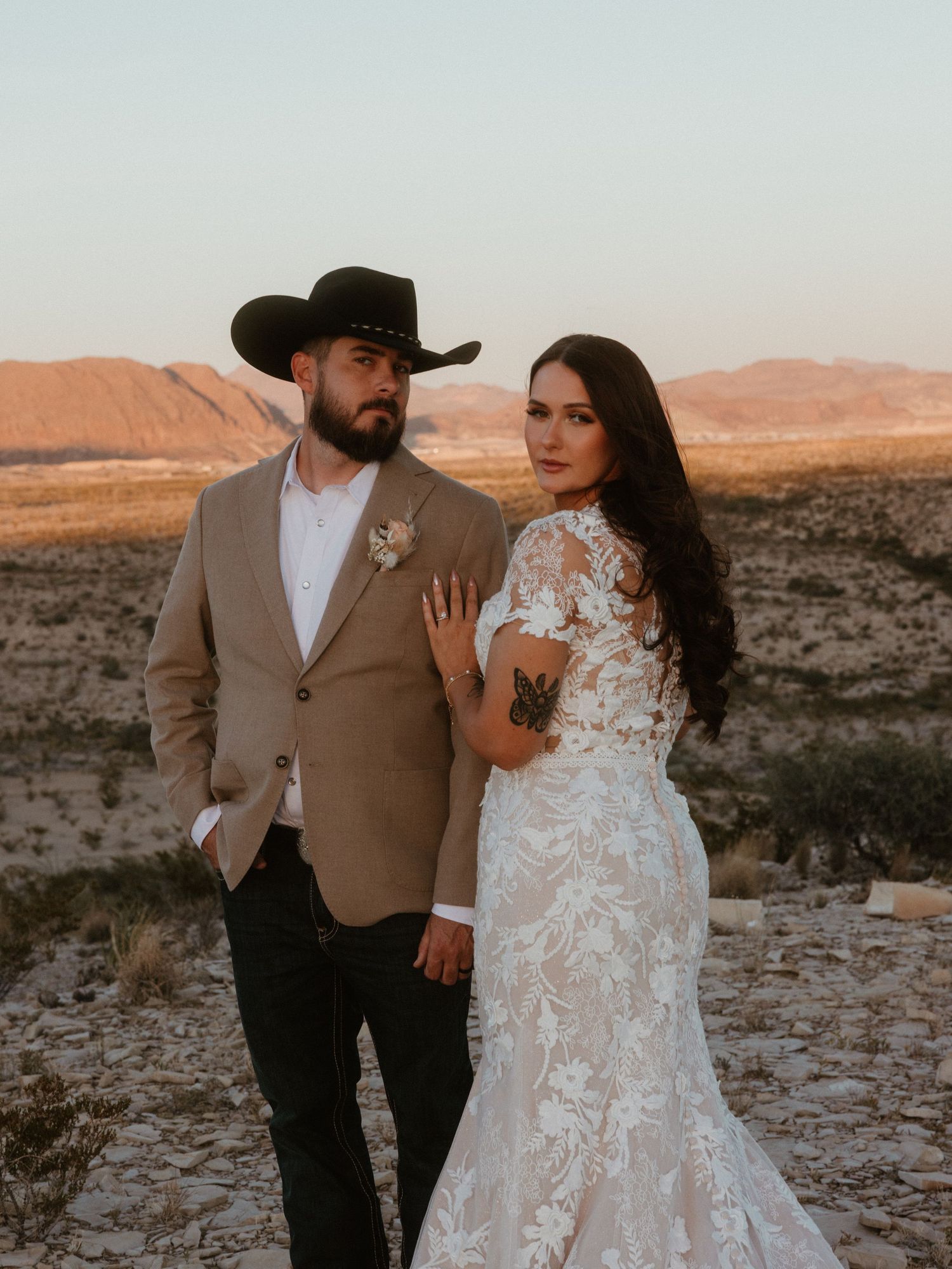 A couple in wedding attire embraces during sunset in a desert landscape with rocky terrain and warm lighting.
