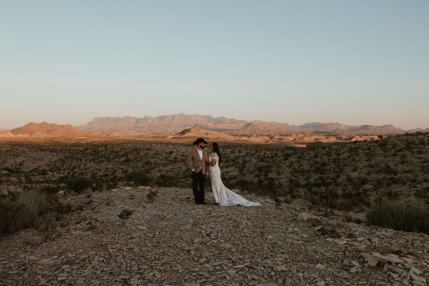 A couple in wedding attire stands in the desert landscape with mountains in the background at sunset.