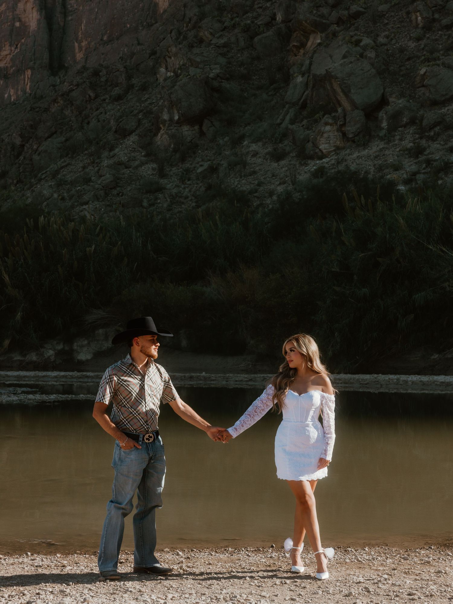 A couple in western and white attire hold hands by a river with dramatic cliffs in the background.