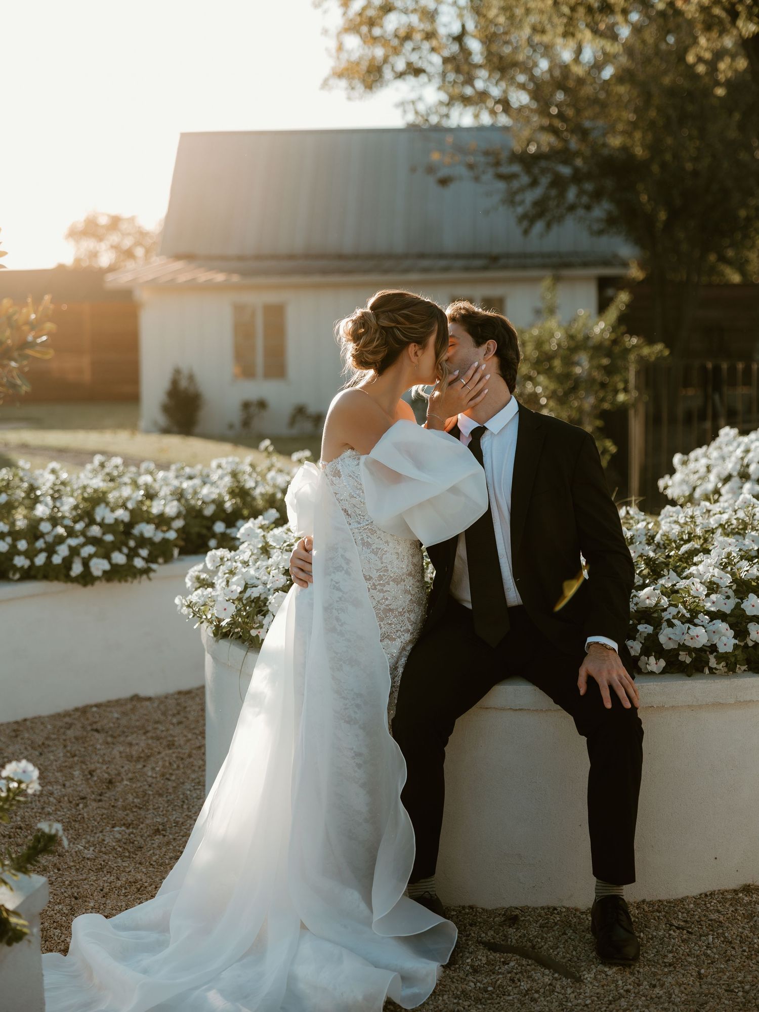 Romantic couple in wedding attire share an intimate moment at sunset among white flowers in a garden setting.