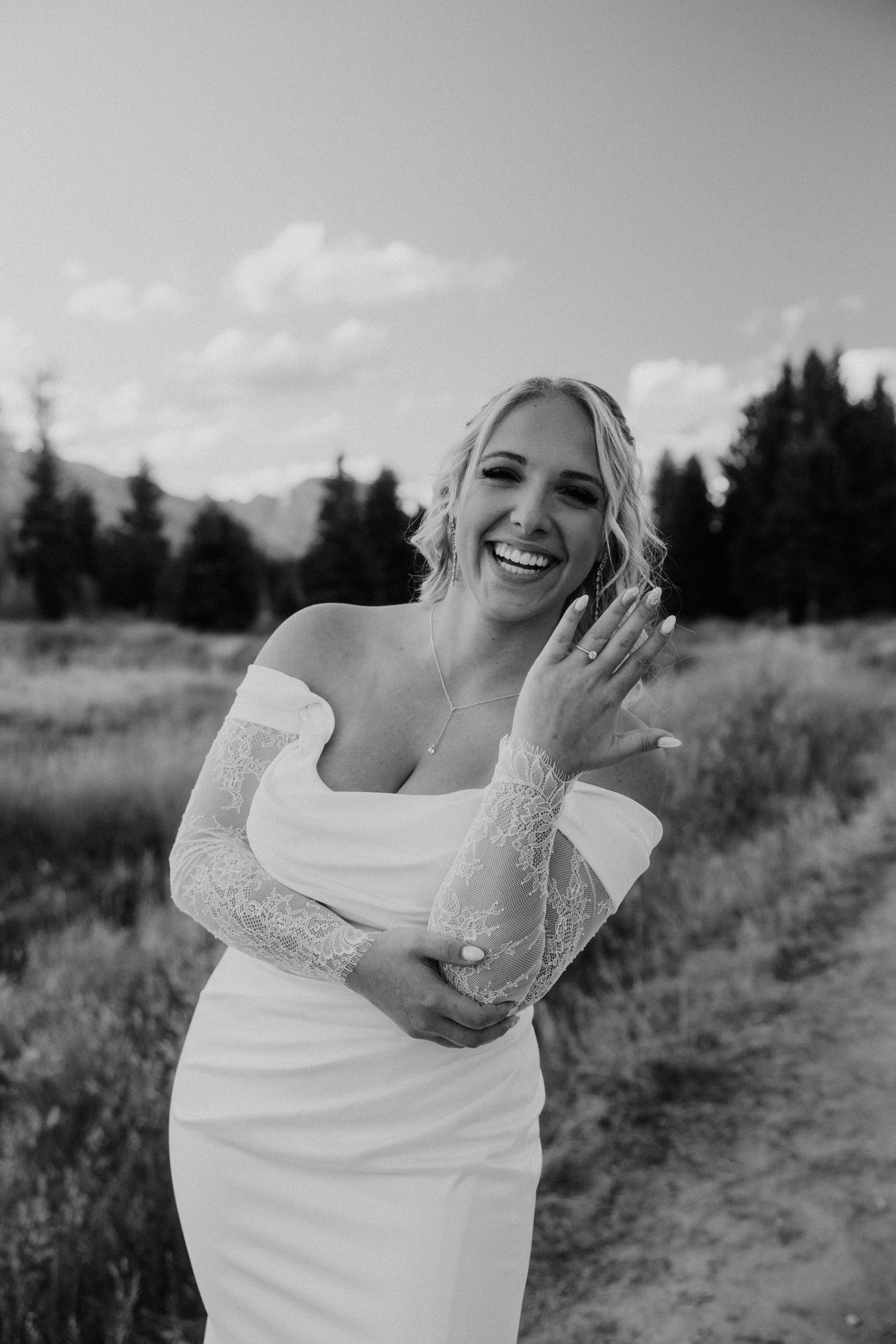 Black and white portrait of bride in lace sleeve dress showing off ring in mountain meadow.