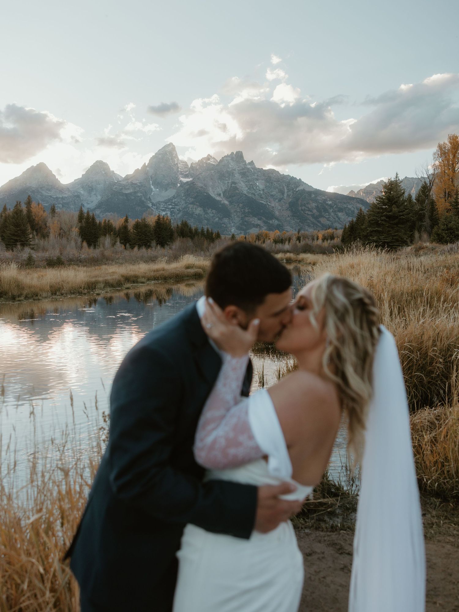 Wedding couple shares kiss with Grand Teton mountains and lake in background at sunset.