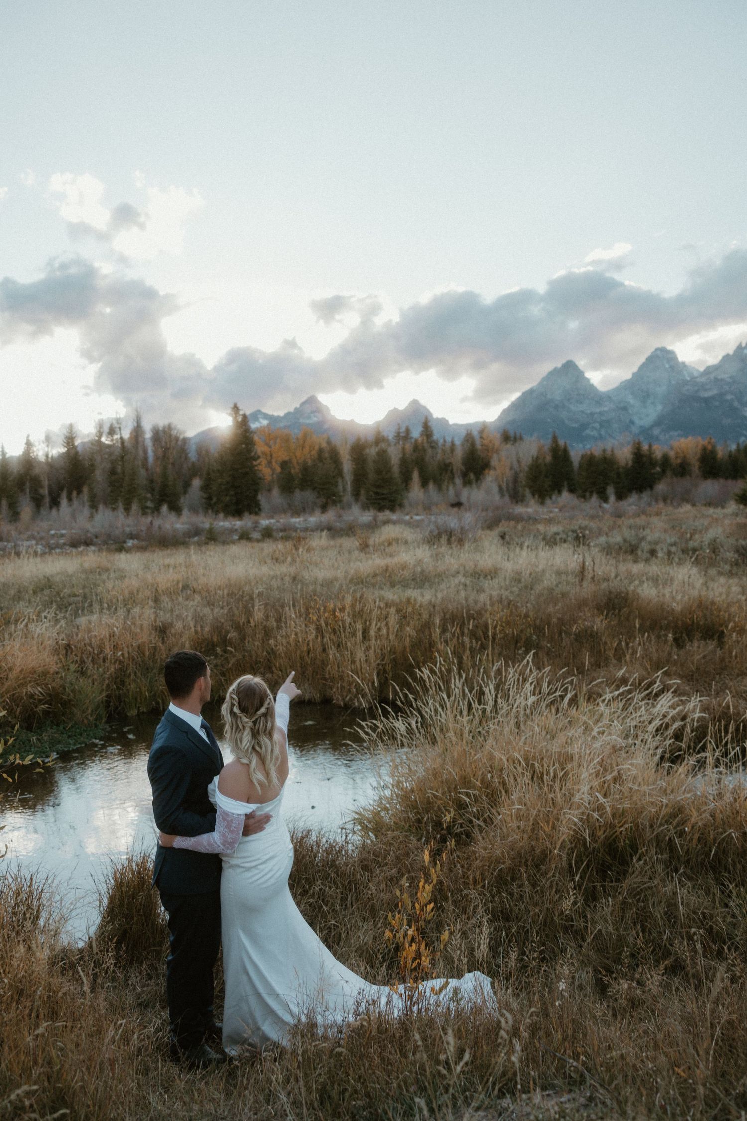 Wedding couple embraces near a lake at sunset with majestic mountains in the distance.