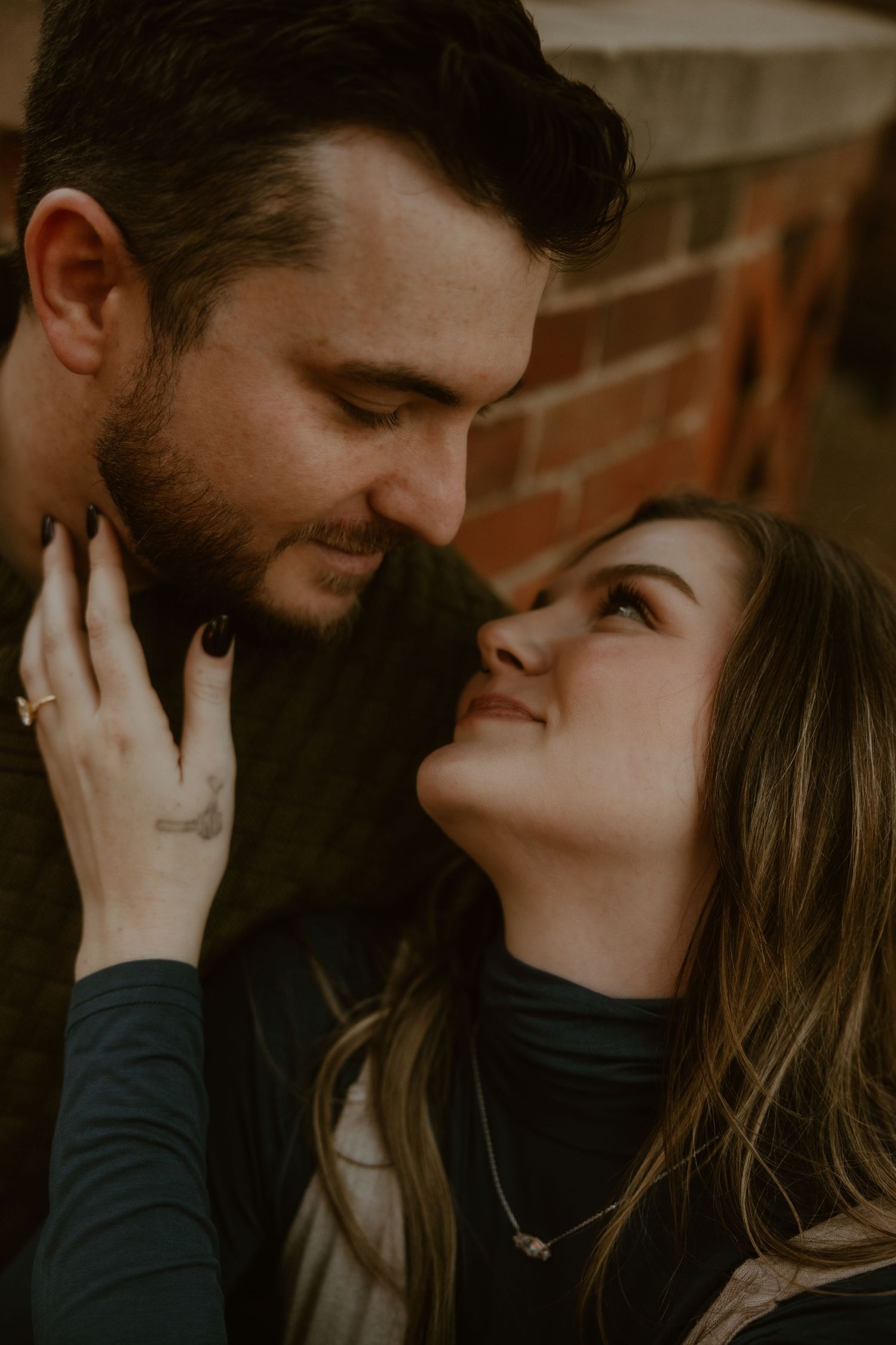 Couple sharing tender moment against brick wall background in moody lighting.