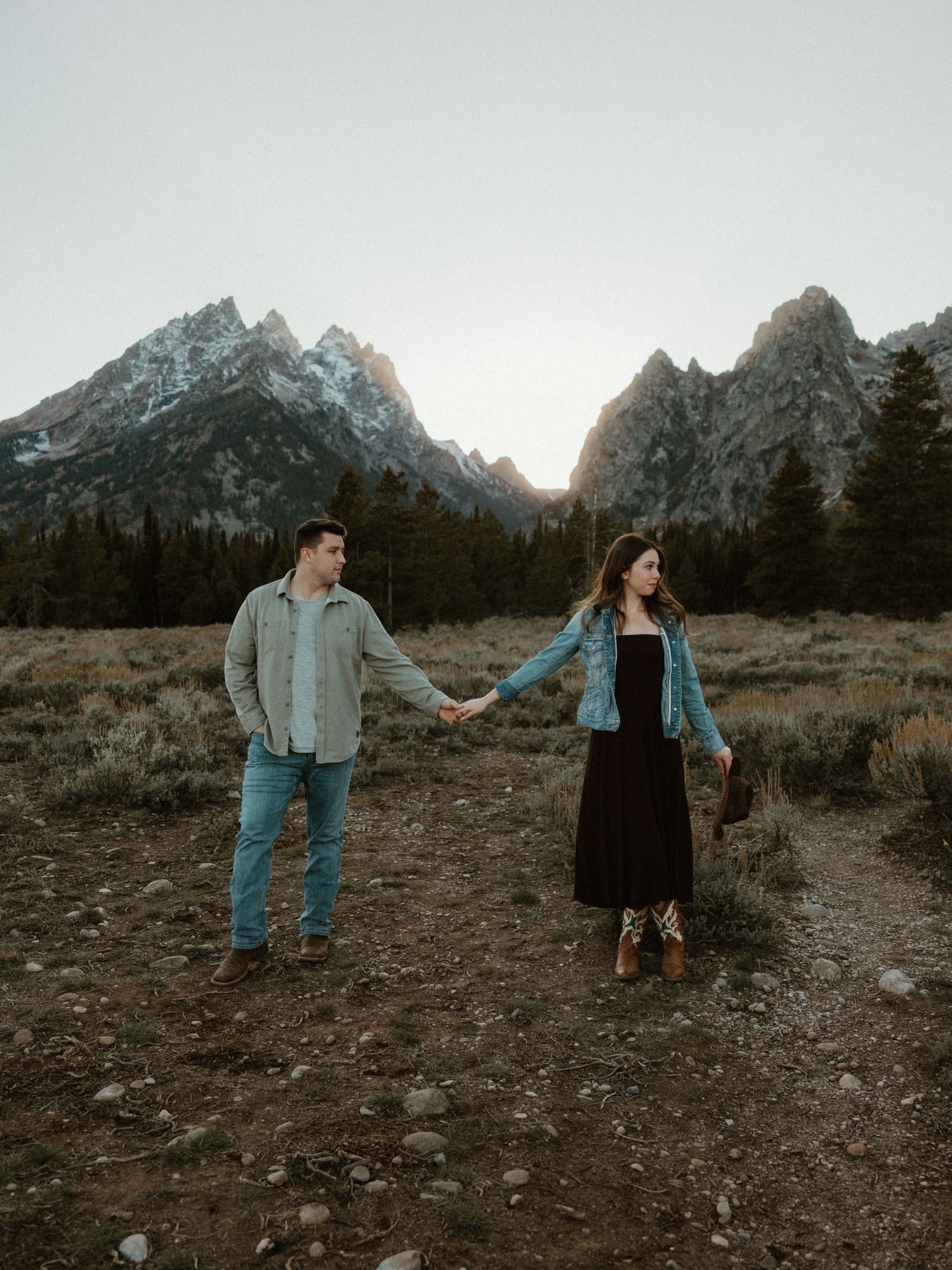 Couple holding hands in front of snow-capped mountain peaks during golden hour.