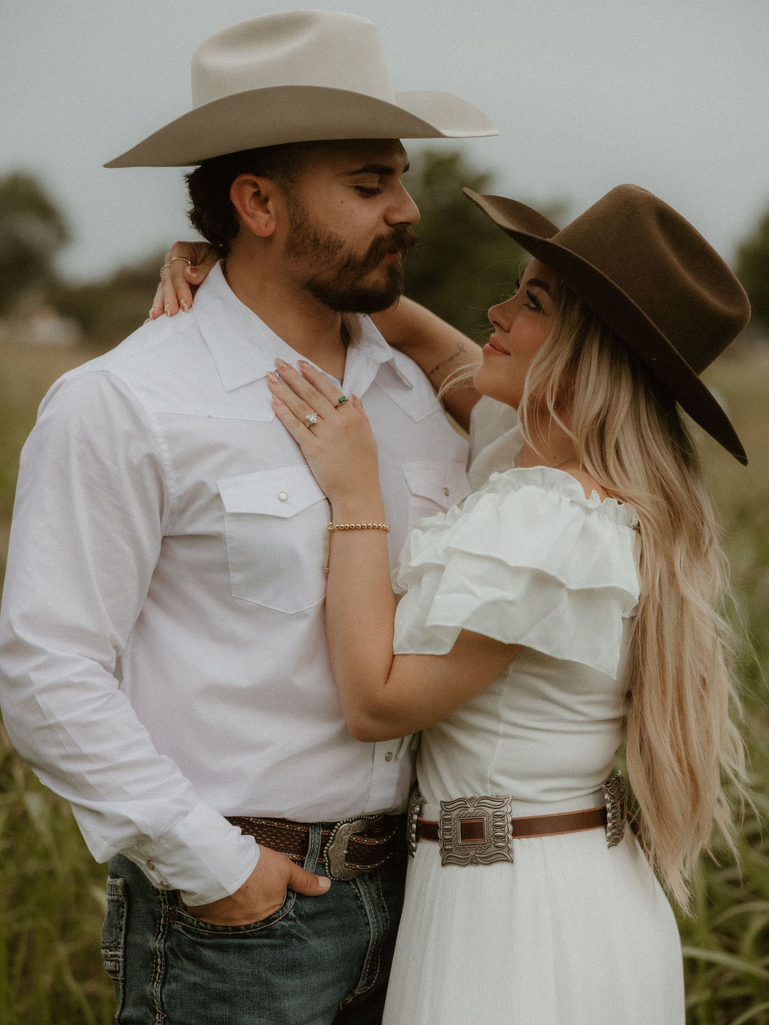 Romantic western-themed couple photoshoot with cowboy hat and white dress in field.