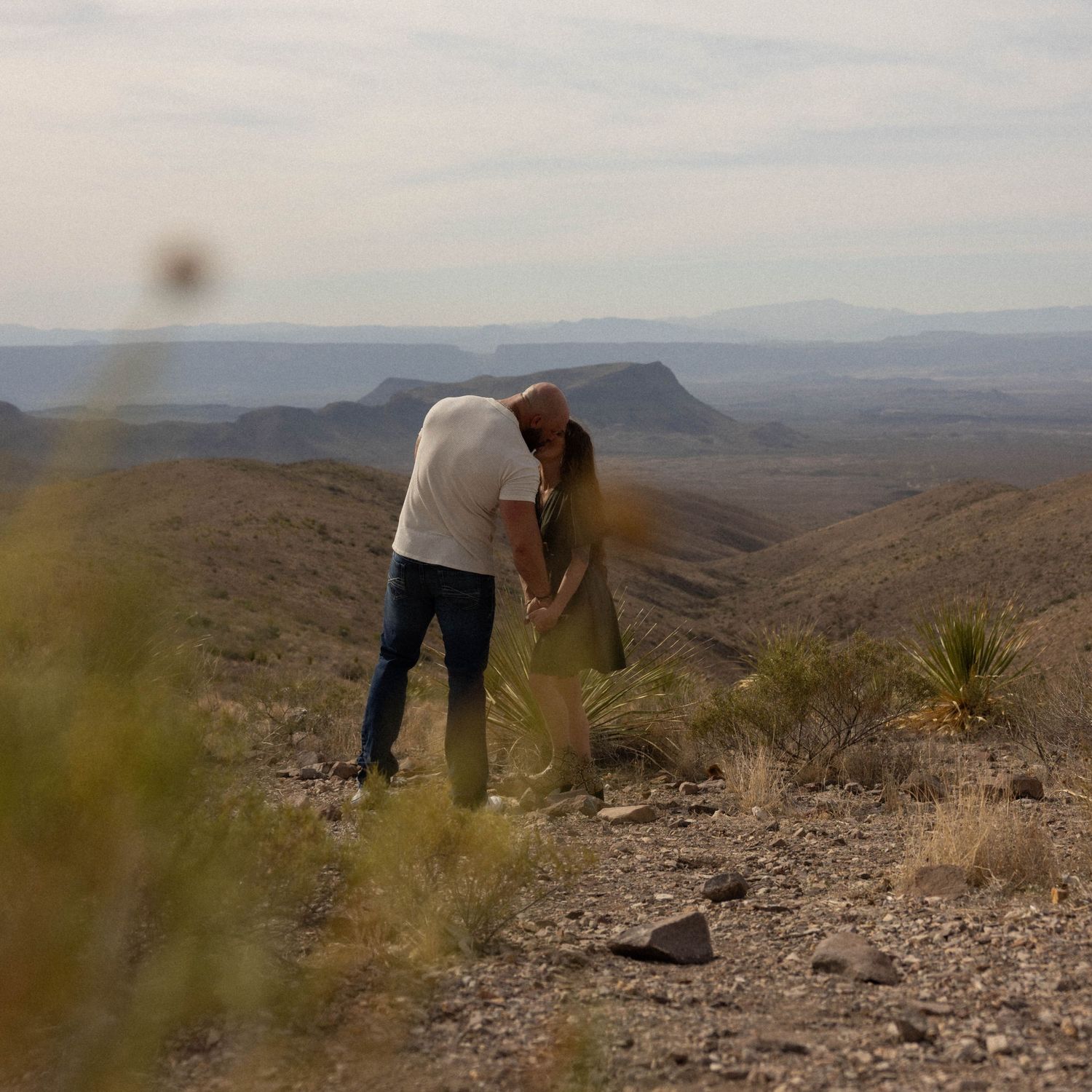 Two figures stand on rocky desert terrain with mountains in the distance.