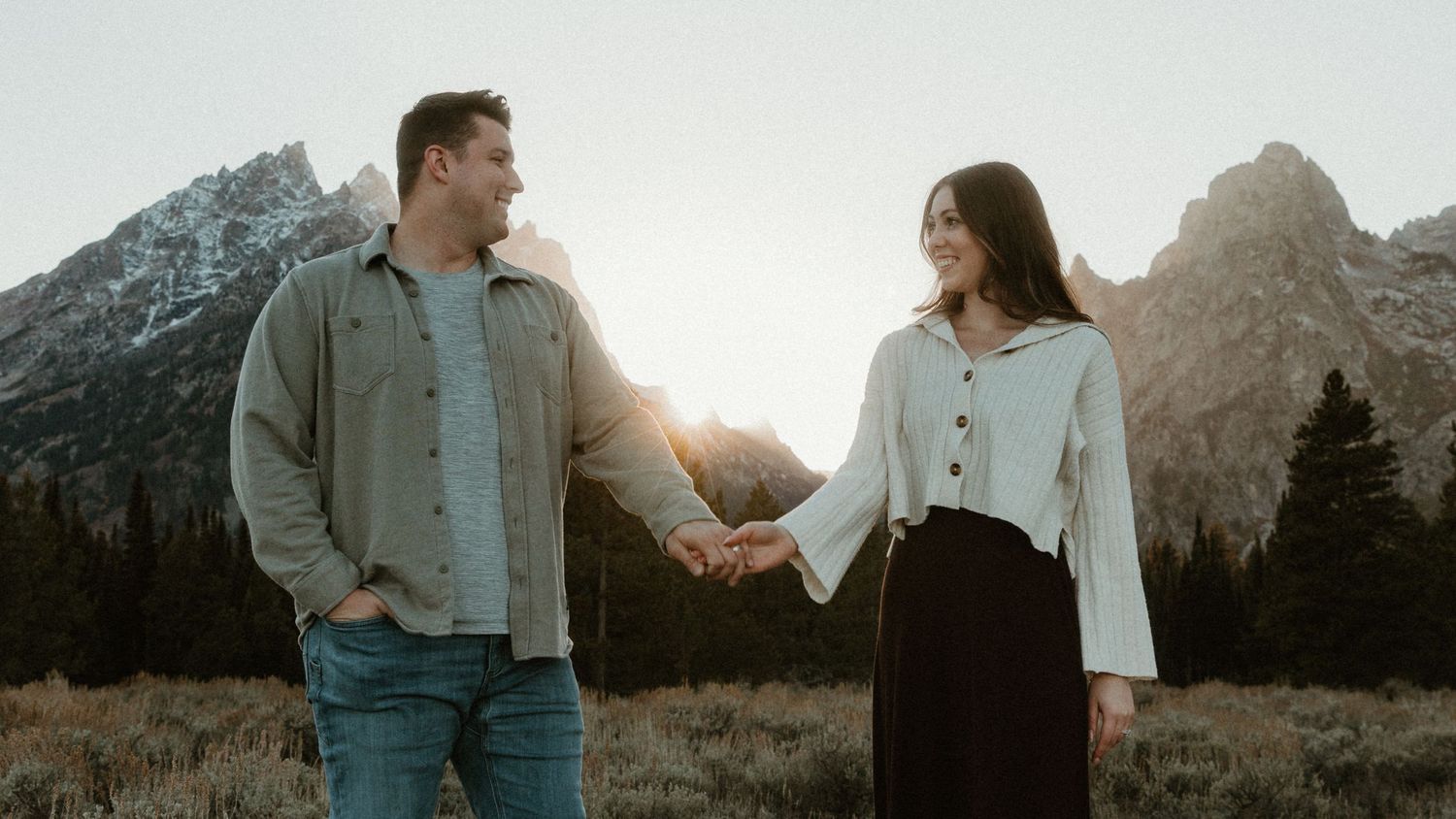 A couple stands together in casual attire with mountain peaks rising behind them.