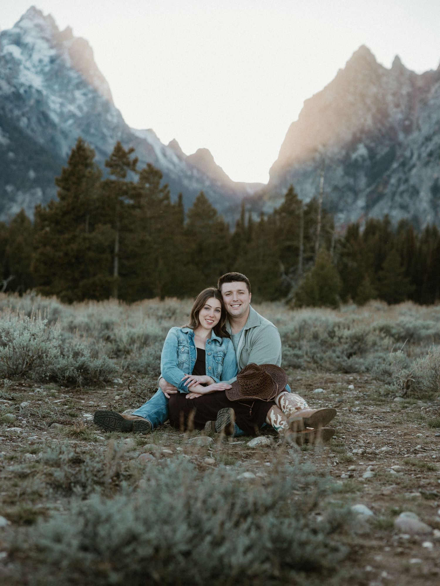 Couple sitting together on ground with Grand Teton mountains in background during sunset.