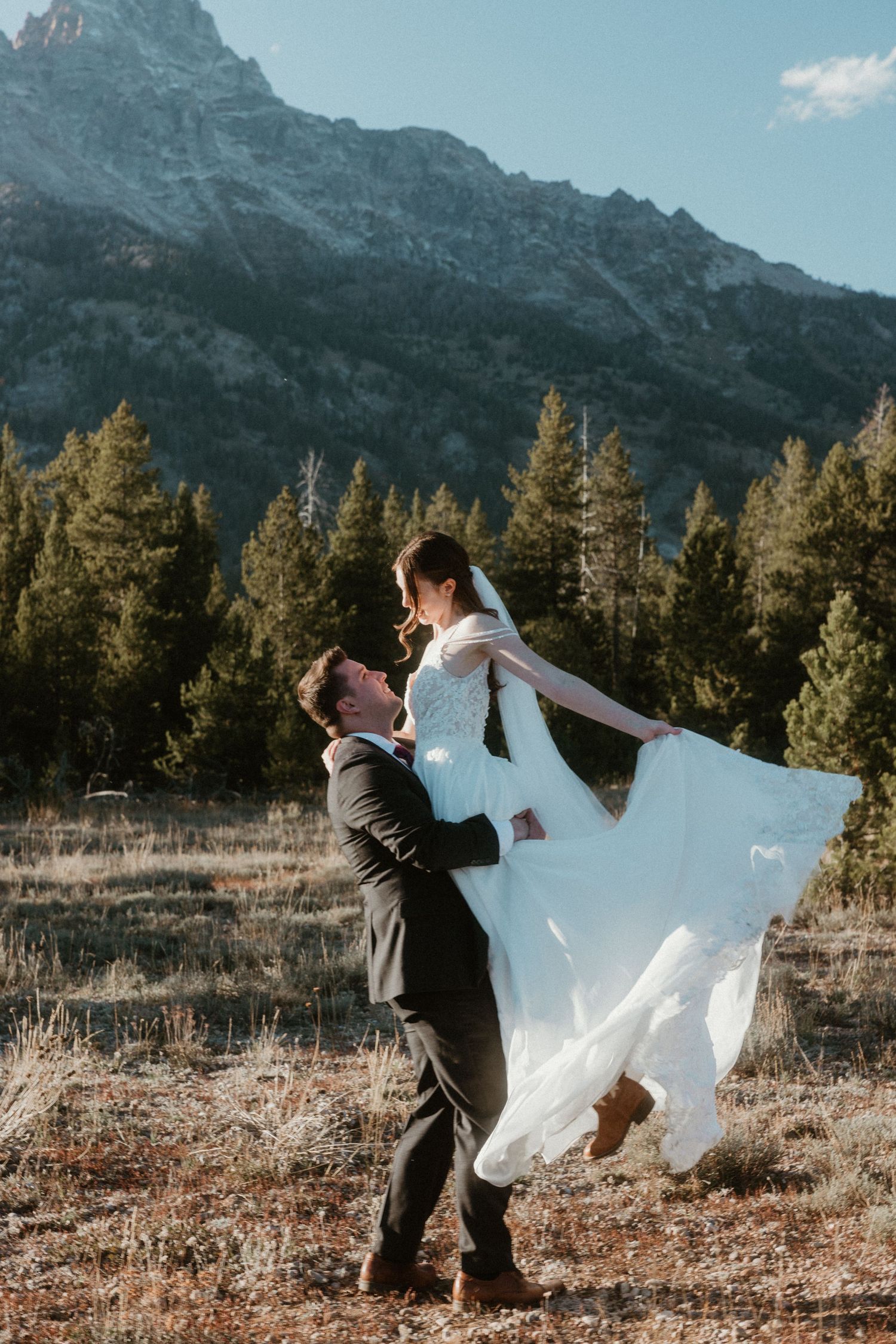 Groom lifts bride in flowing white dress against mountain landscape.