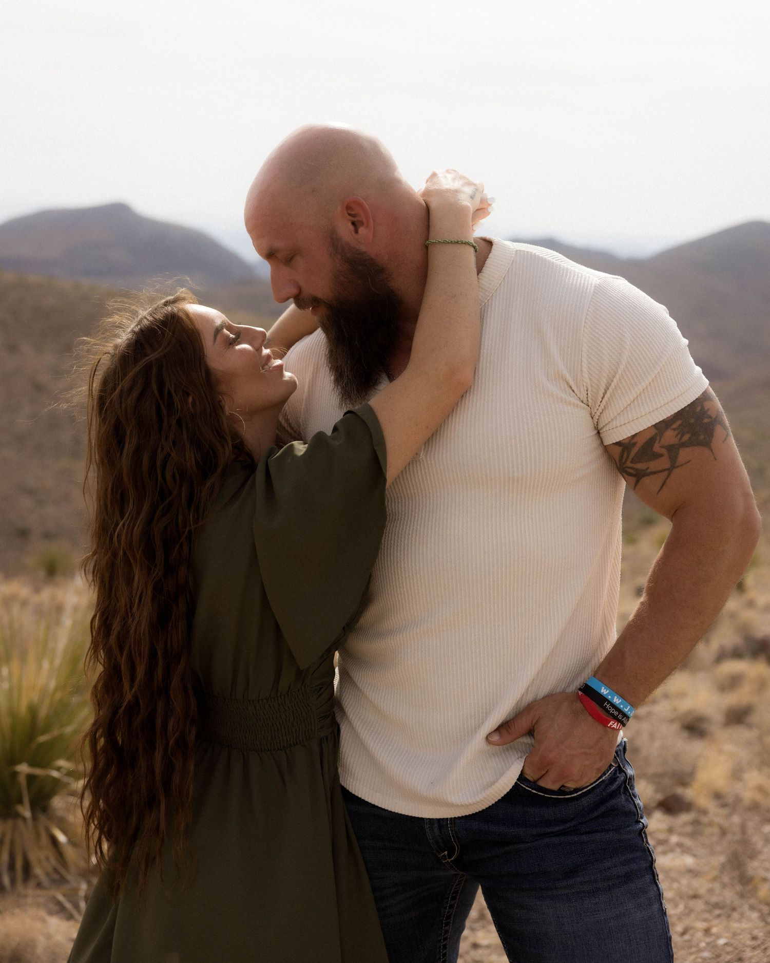 A couple embraces while standing in a desert landscape with mountains behind them.