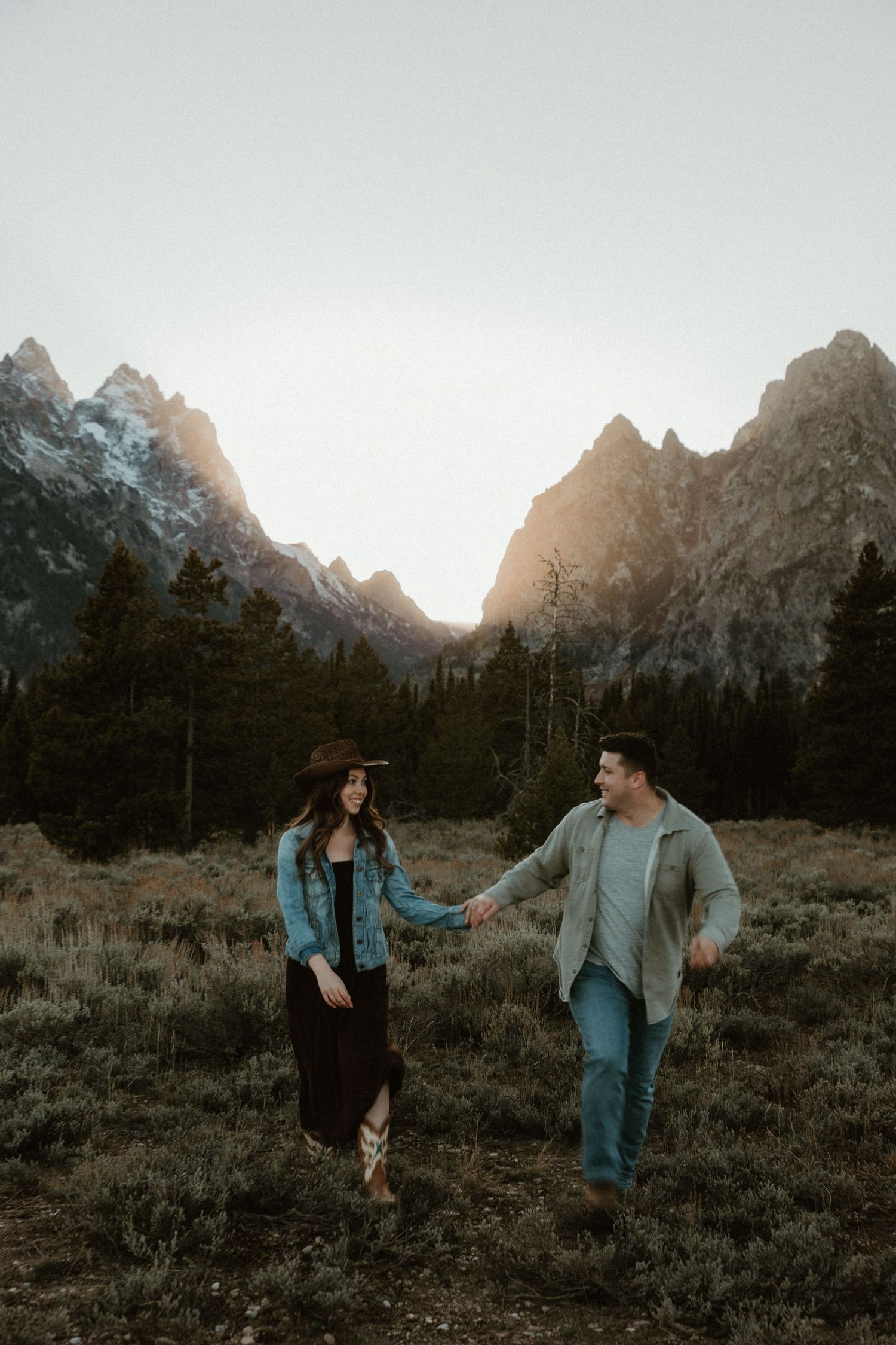 Playful couple in denim and grey walking through mountain meadow at sunset.