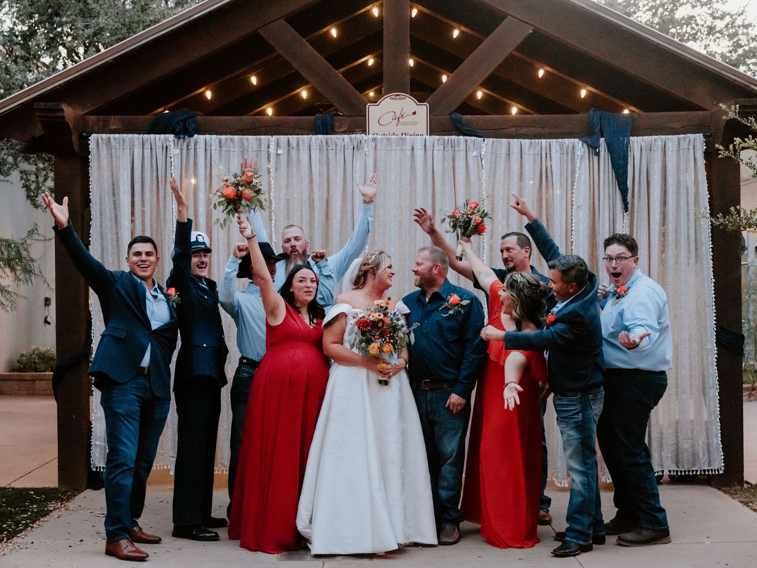 Wedding party celebrates with raised arms outside rustic venue with string lights.