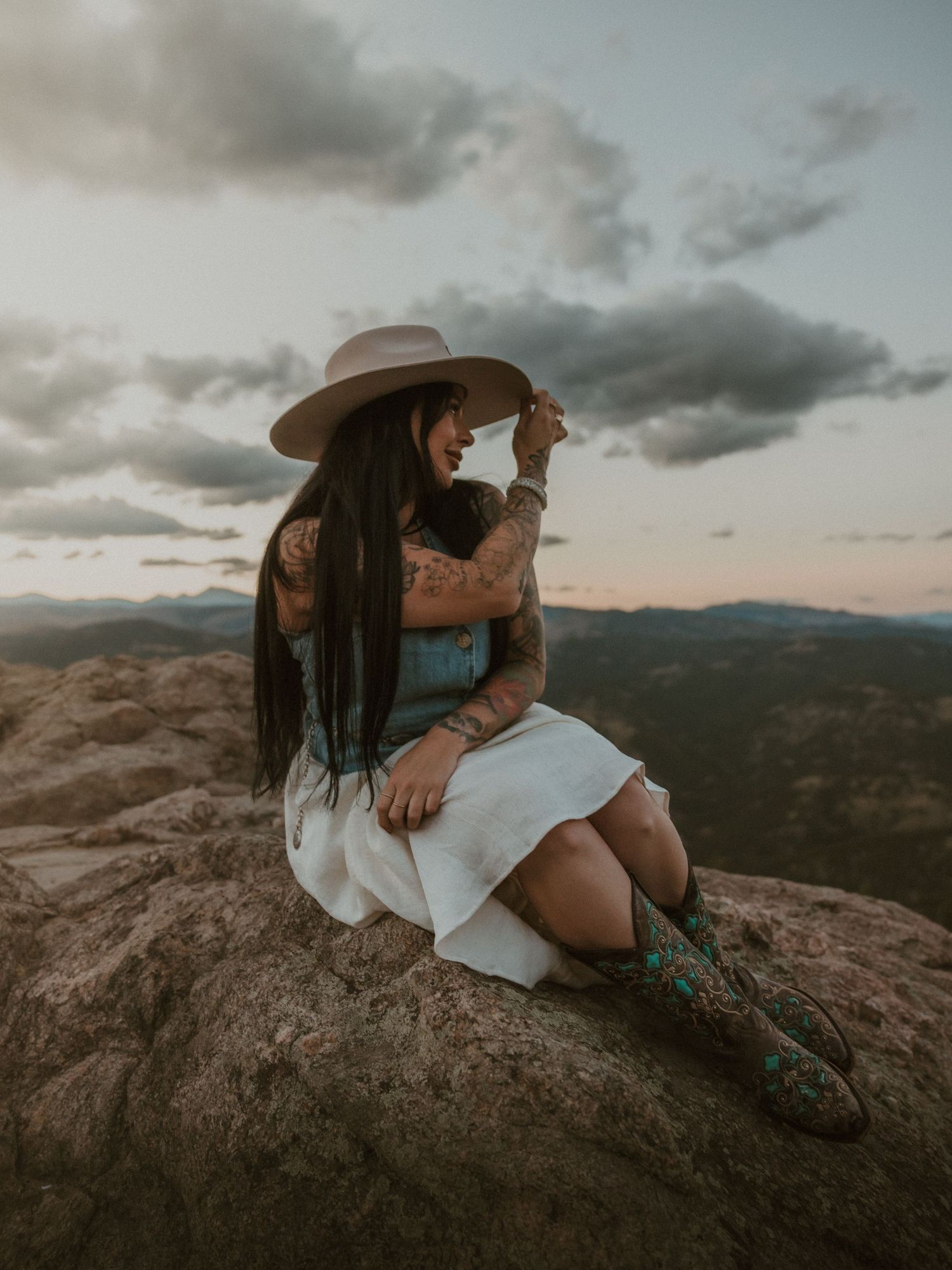 Person in a hat and white dress sits on mountain peak at sunset with dramatic cloudy sky in background.