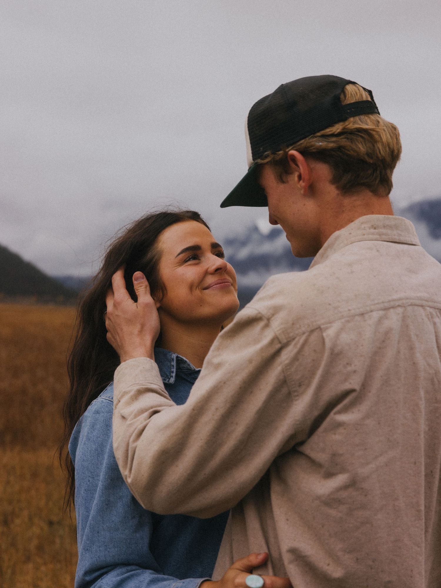 A romantic couple embraces in a field with mountains in the background during golden hour.