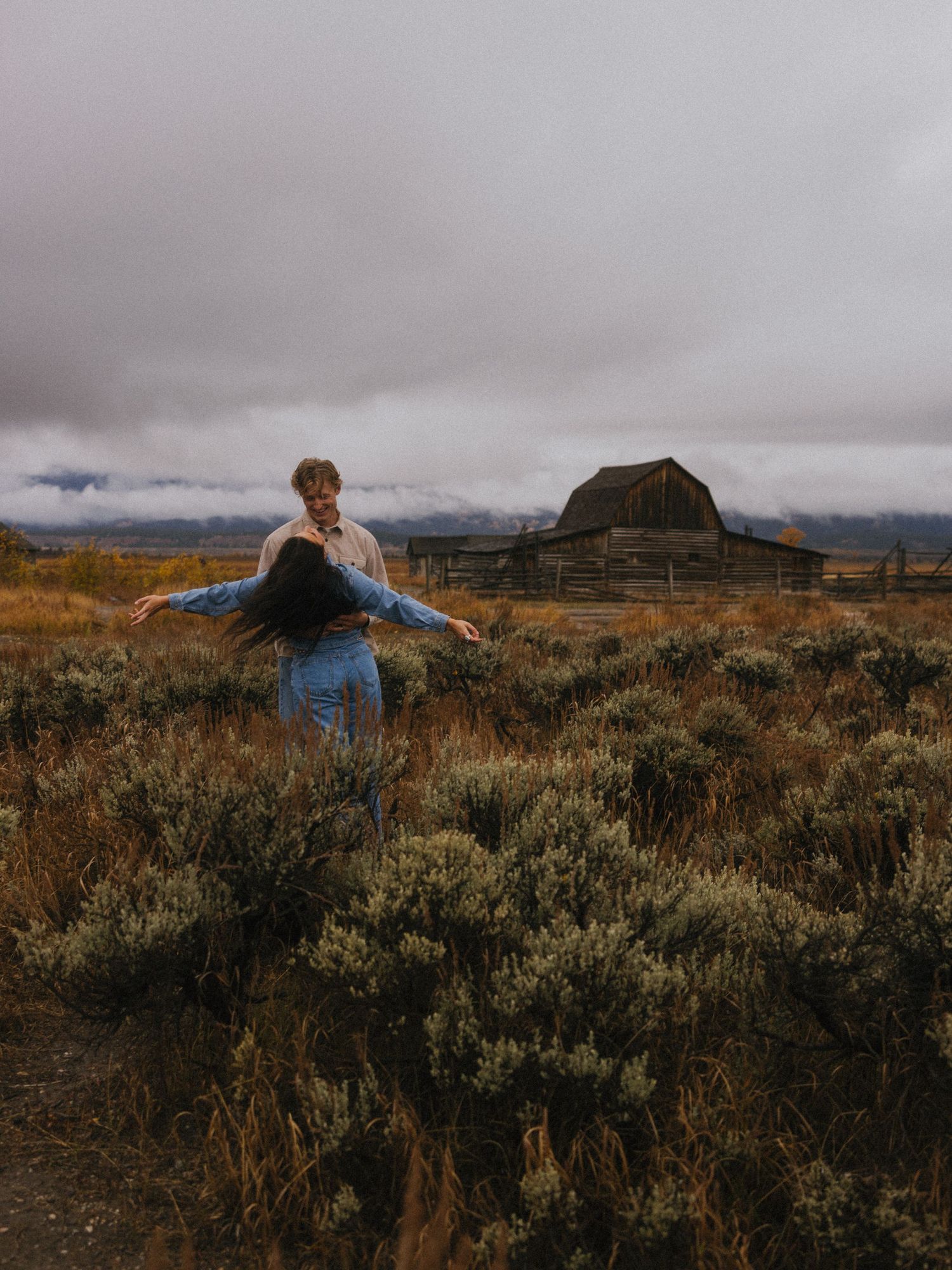 Two people share a playful moment in a rustic field with mountains in the background.