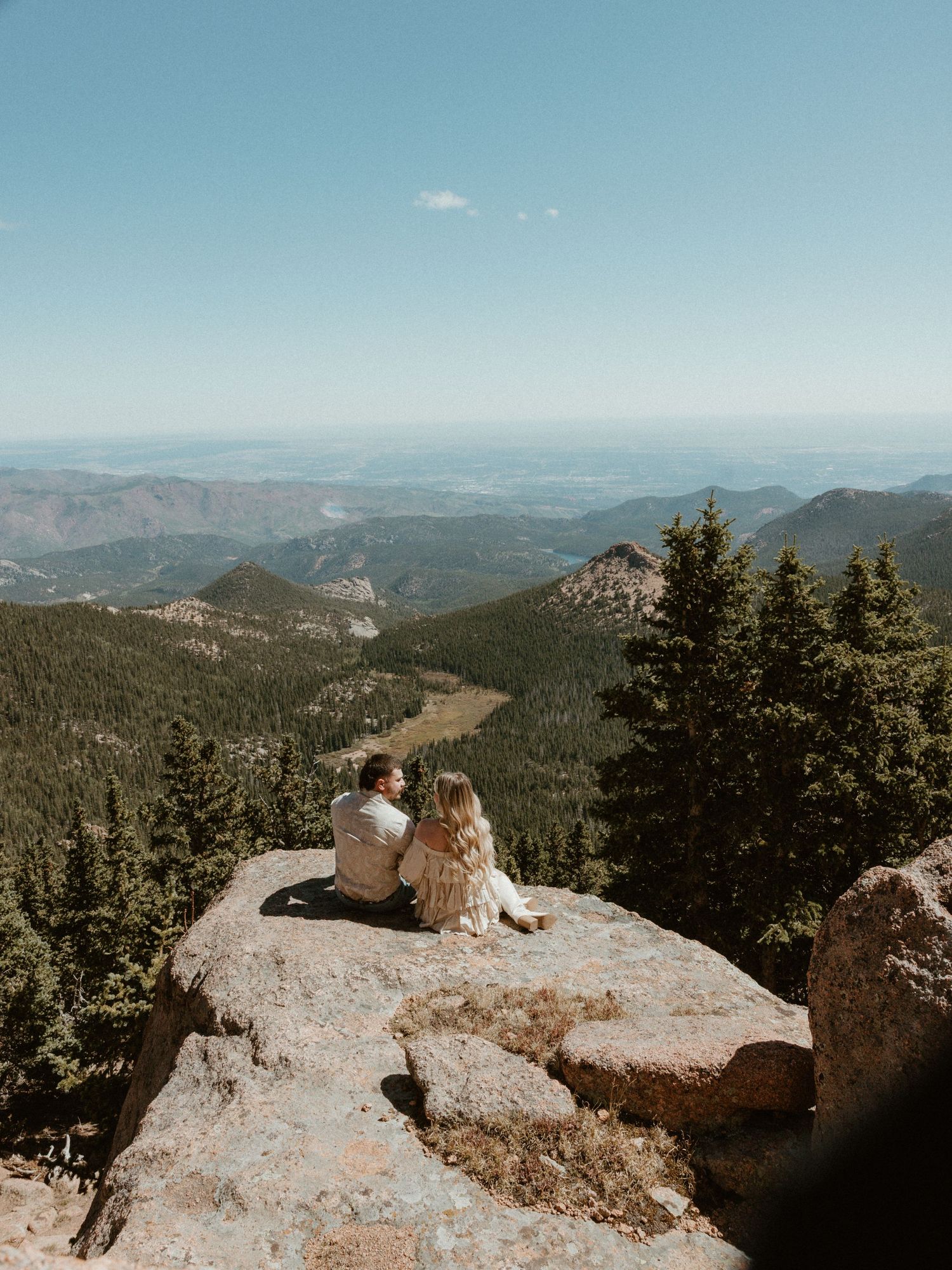 Two people sit close together on mountain ledge enjoying panoramic forest and mountain views.