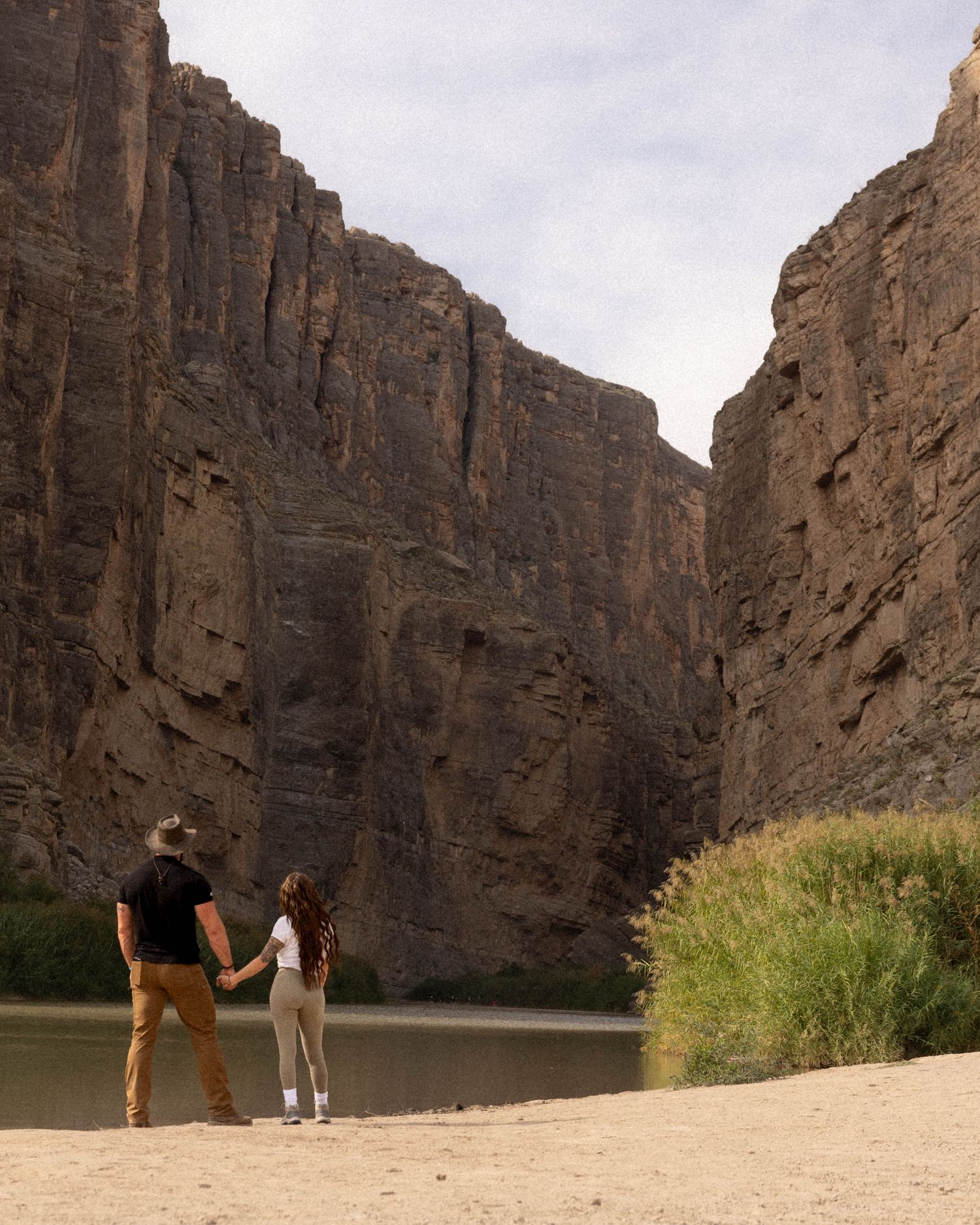 Tall canyon walls frame a sandy riverbank with green vegetation along the water's edge.
