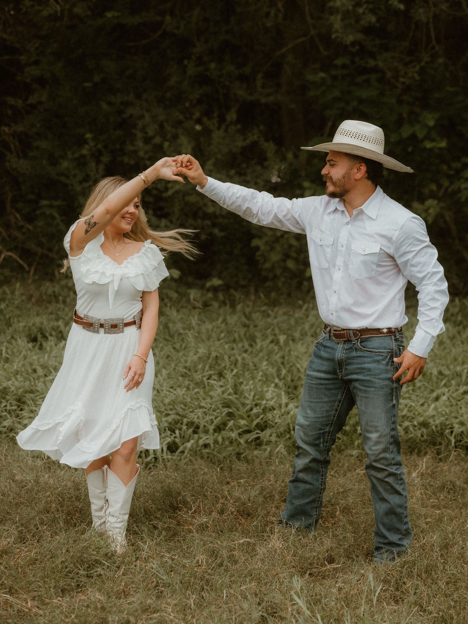 Playful couple in white and denim dancing in a rustic field setting.