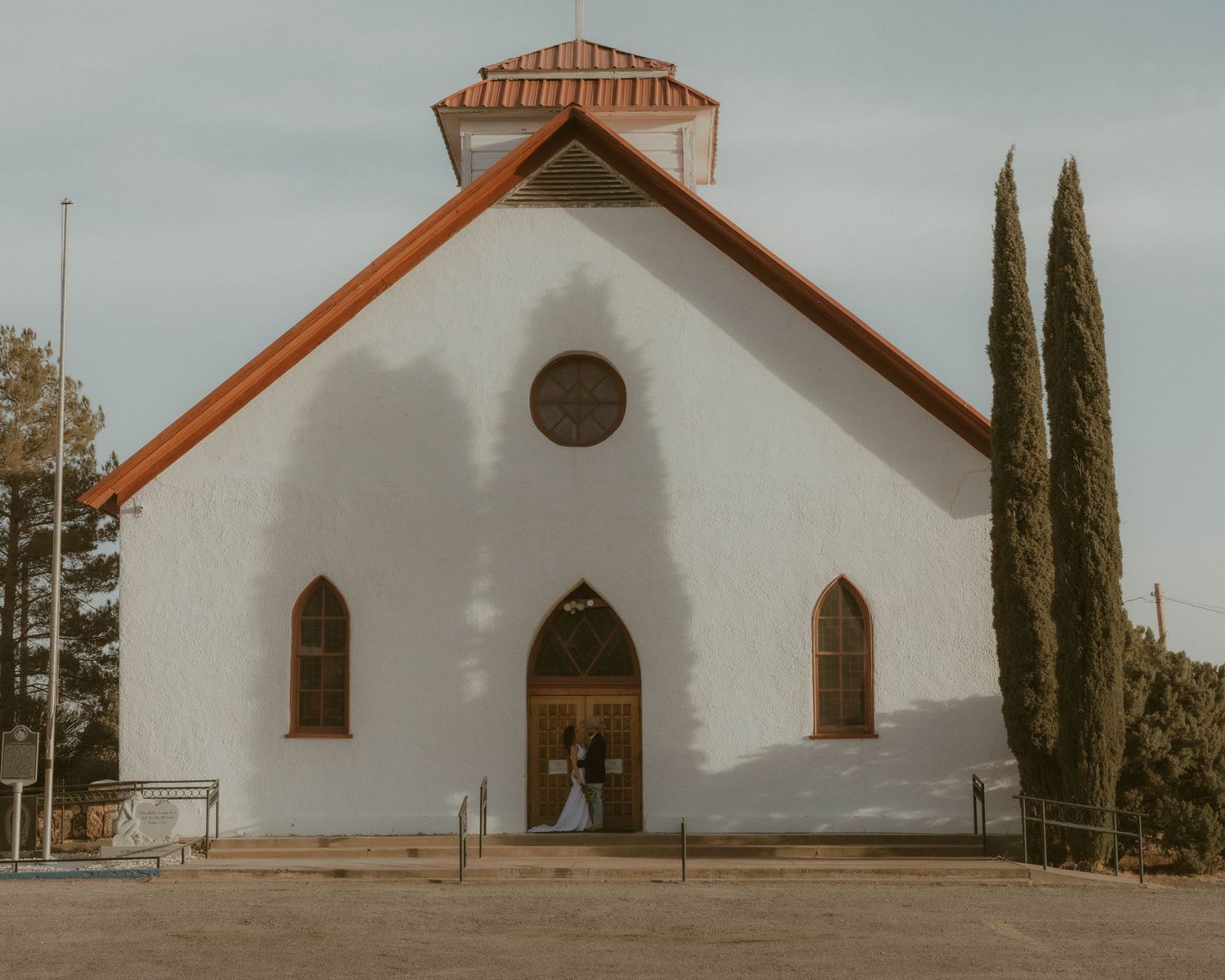 White church exterior with Spanish-style architecture and cypress trees against a muted sky.