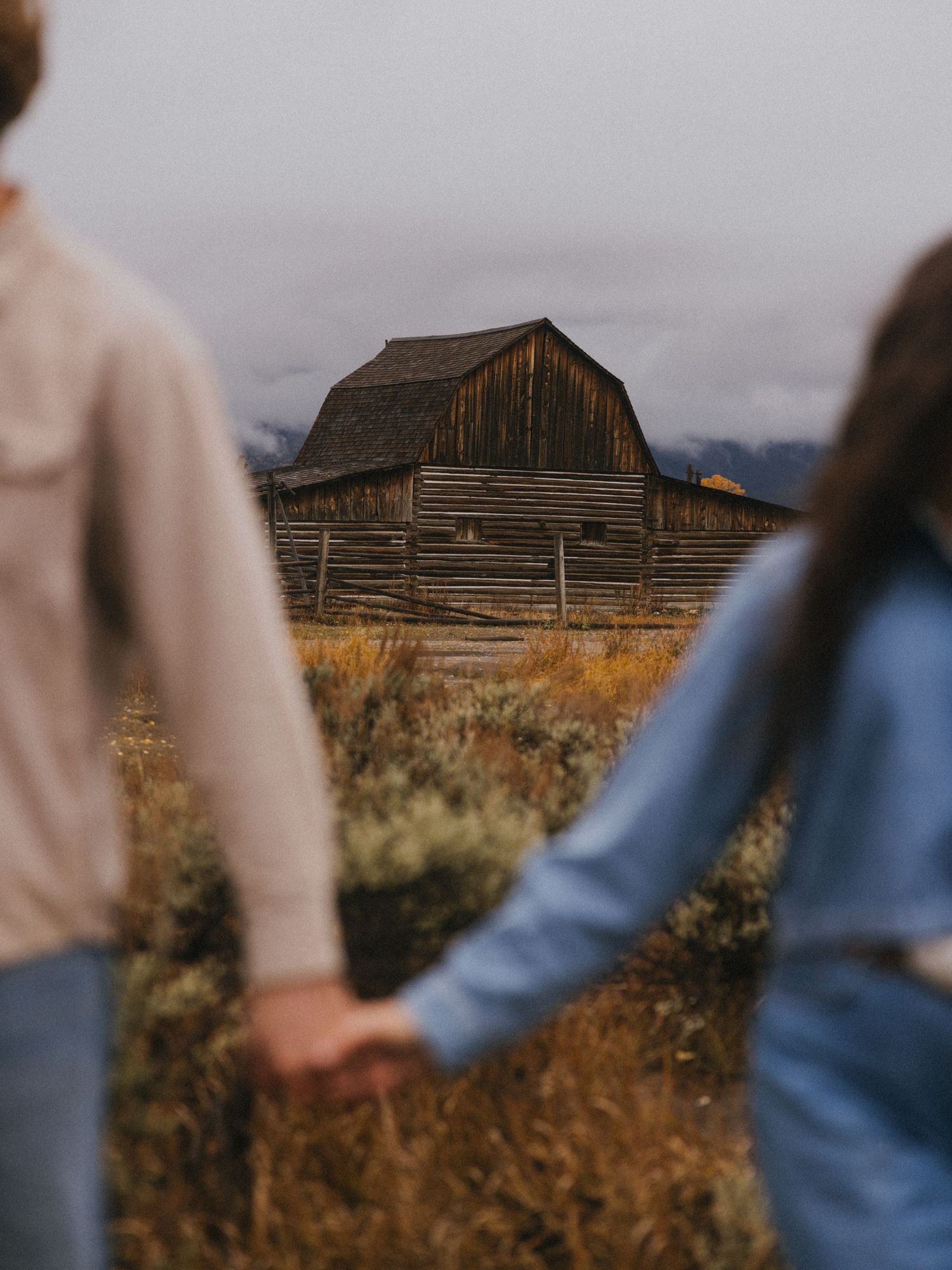 Two people stroll through autumn grass with an old wooden barn in the background.