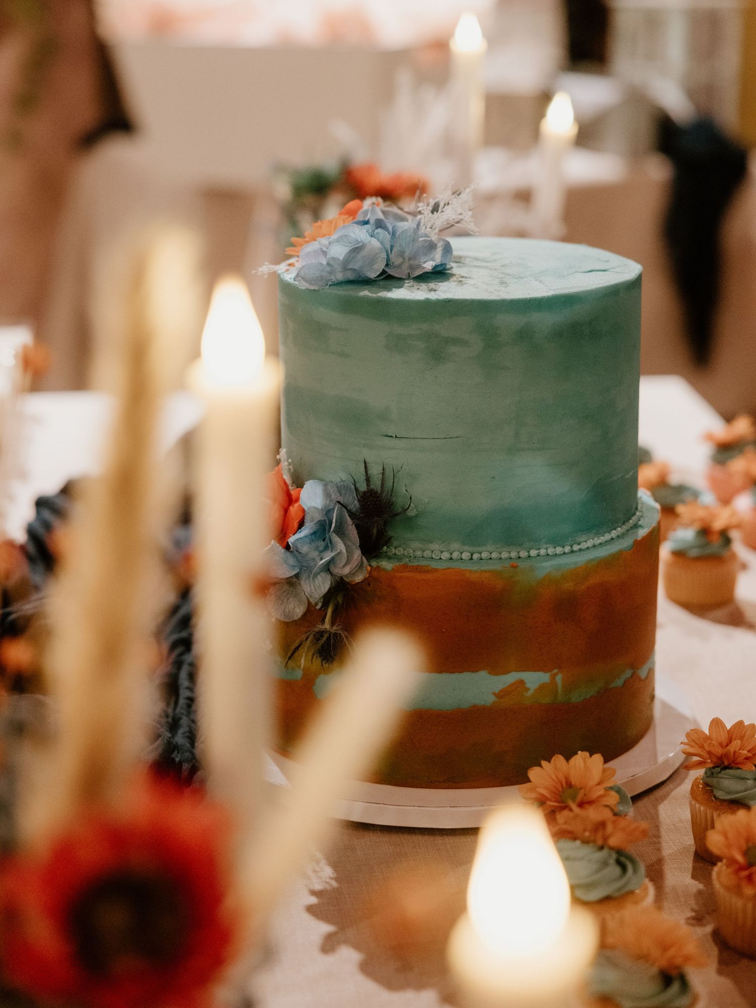A two-tiered wedding cake decorated in teal and copper colors with floral accents and lit candles.