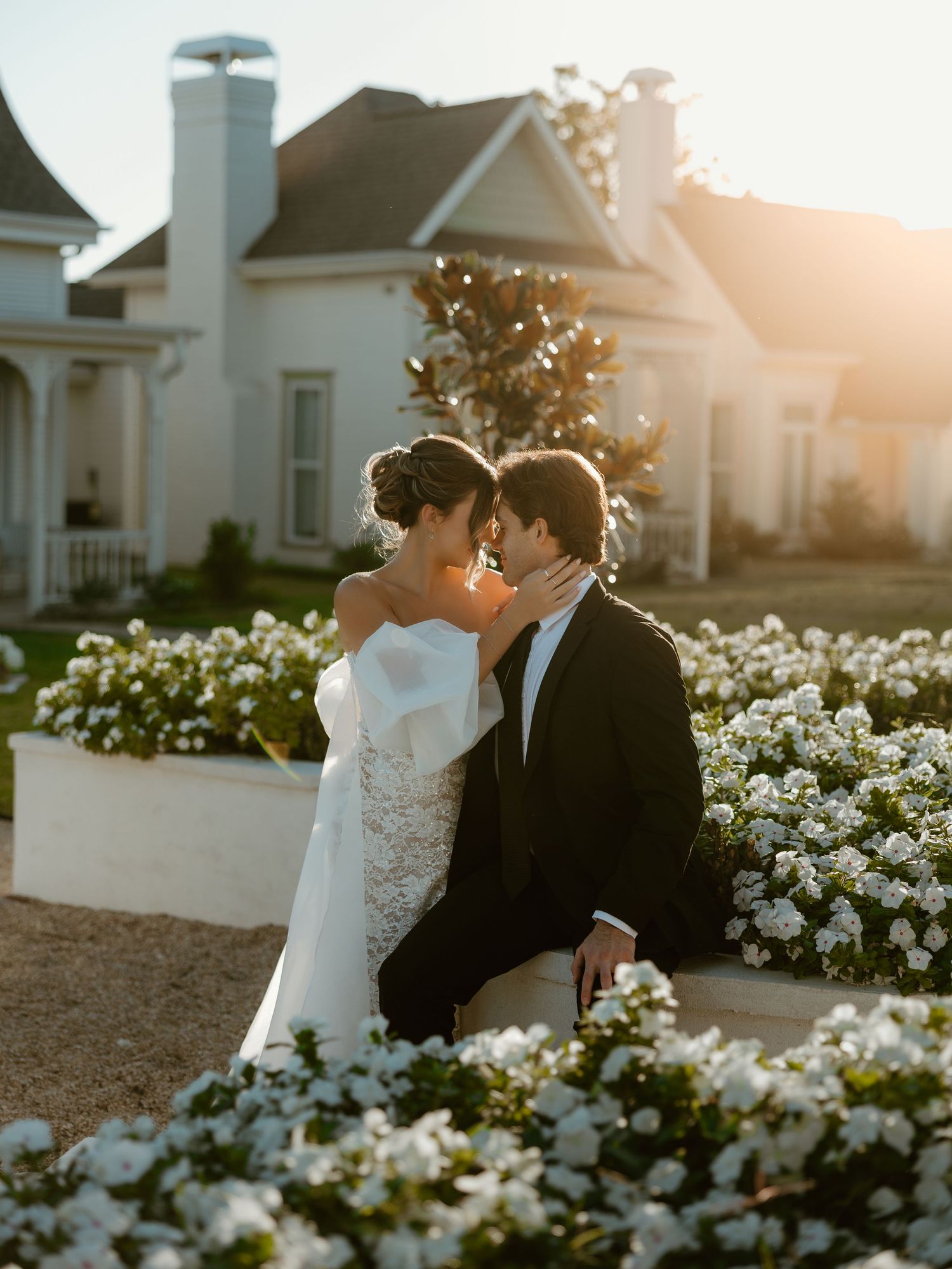 A couple shares an intimate moment among blooming white flowers during golden hour sunset.