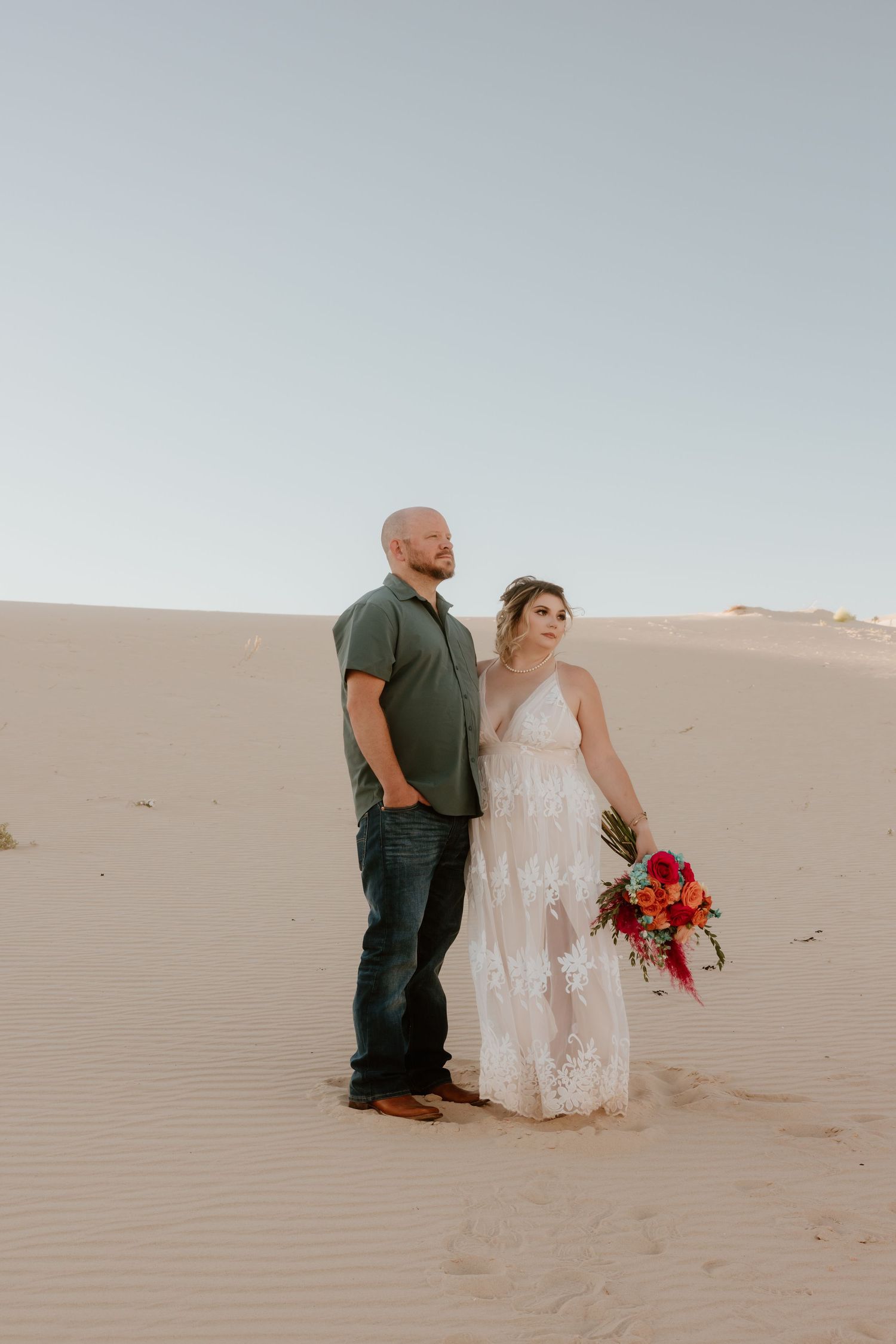 A couple poses together in a desert setting with the bride holding a vibrant red bouquet.