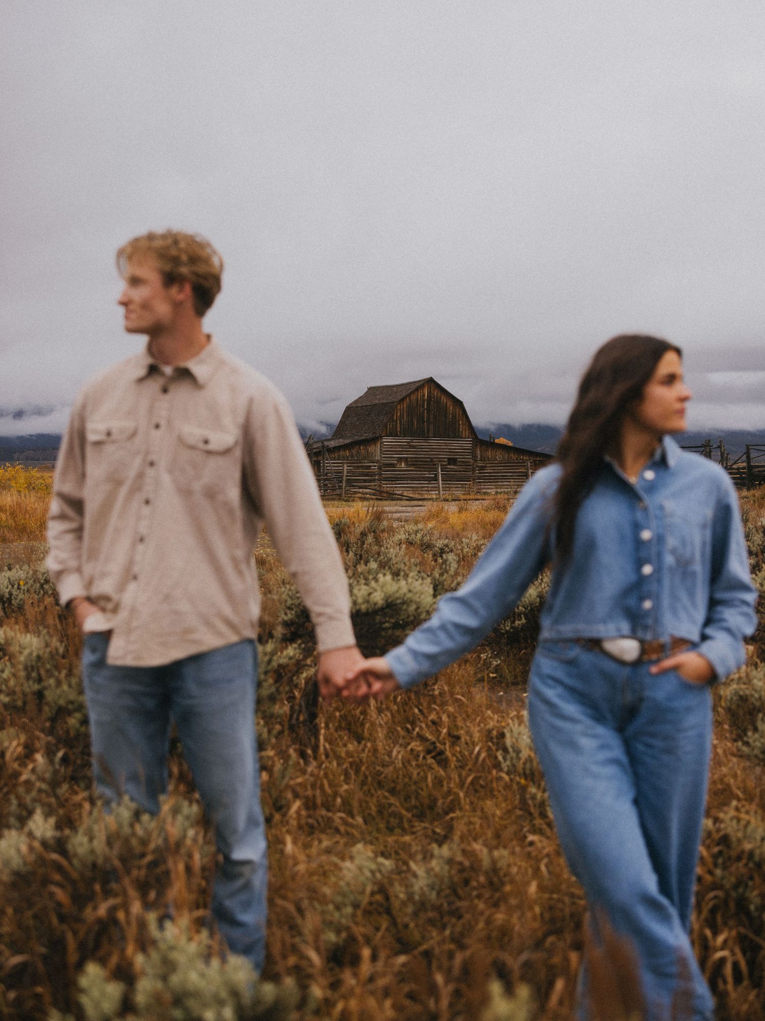 A couple in casual denim and beige attire walks through a field near rustic buildings.