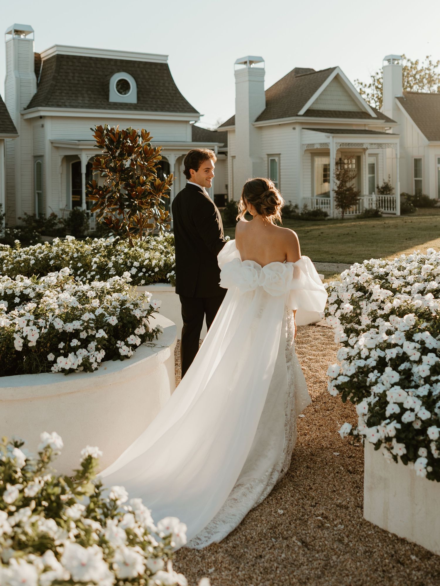 A romantic wedding moment captured in a garden path lined with blooming white flowers at sunset.