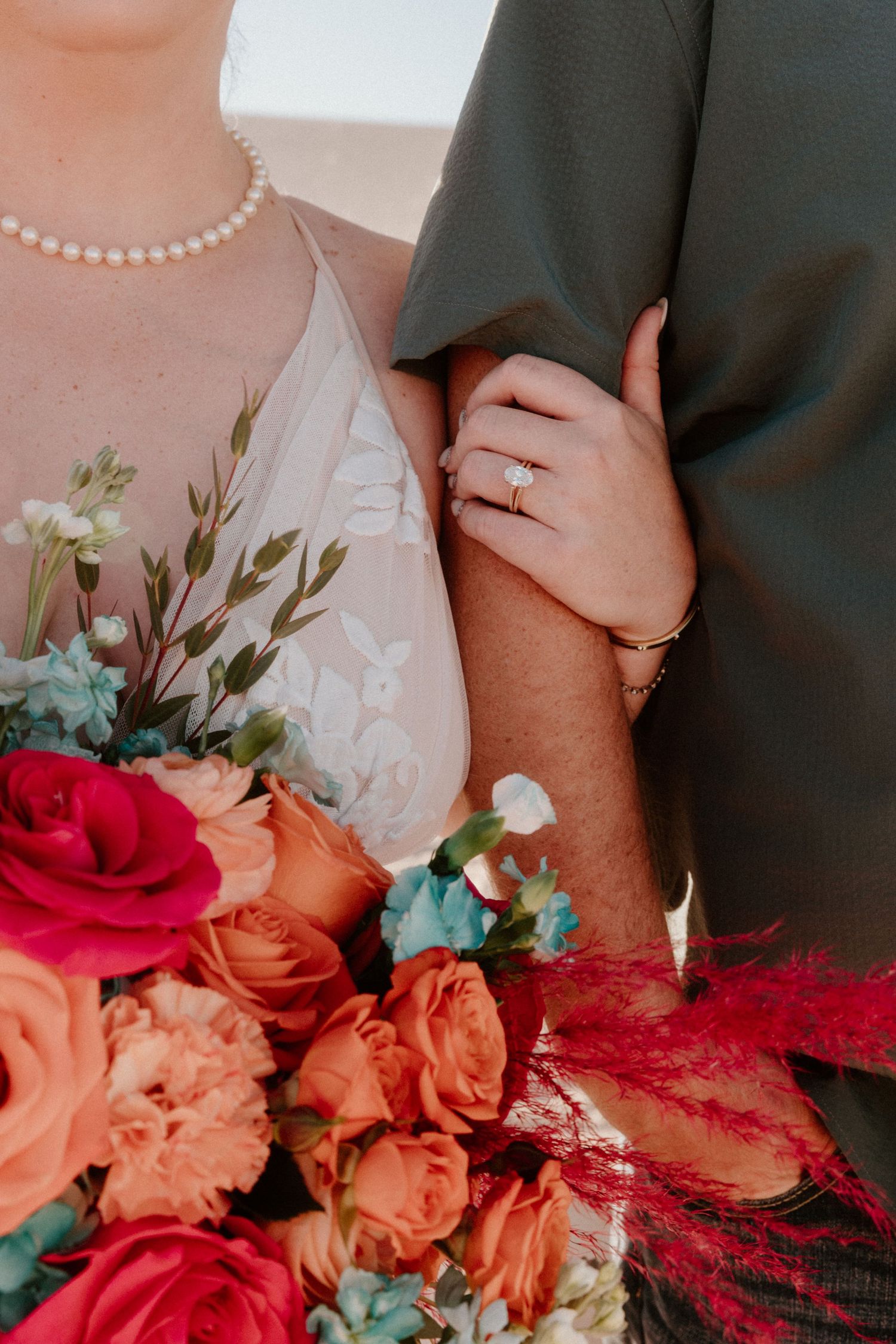 Wedding detail shot of couple's hands with vibrant red and coral roses in foreground.