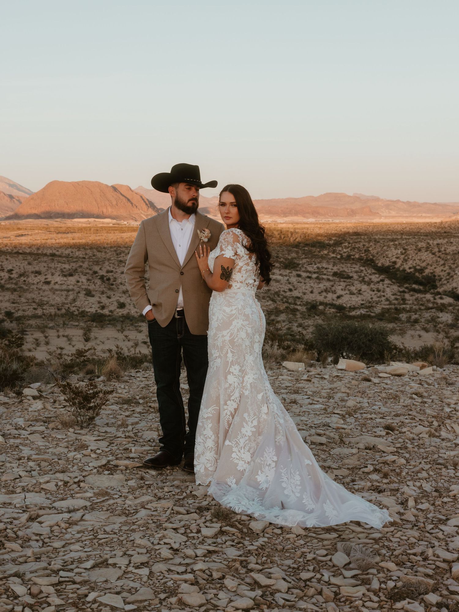 A couple in wedding attire poses on rocky desert terrain at sunset.
