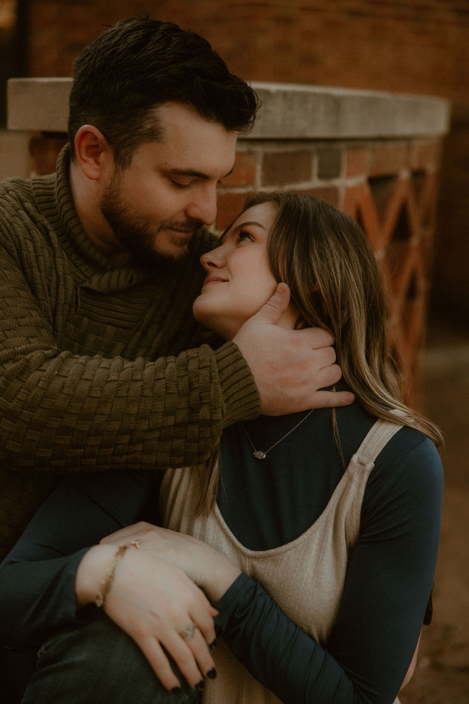 Couple in cozy sweaters sharing intimate moment against brick wall.