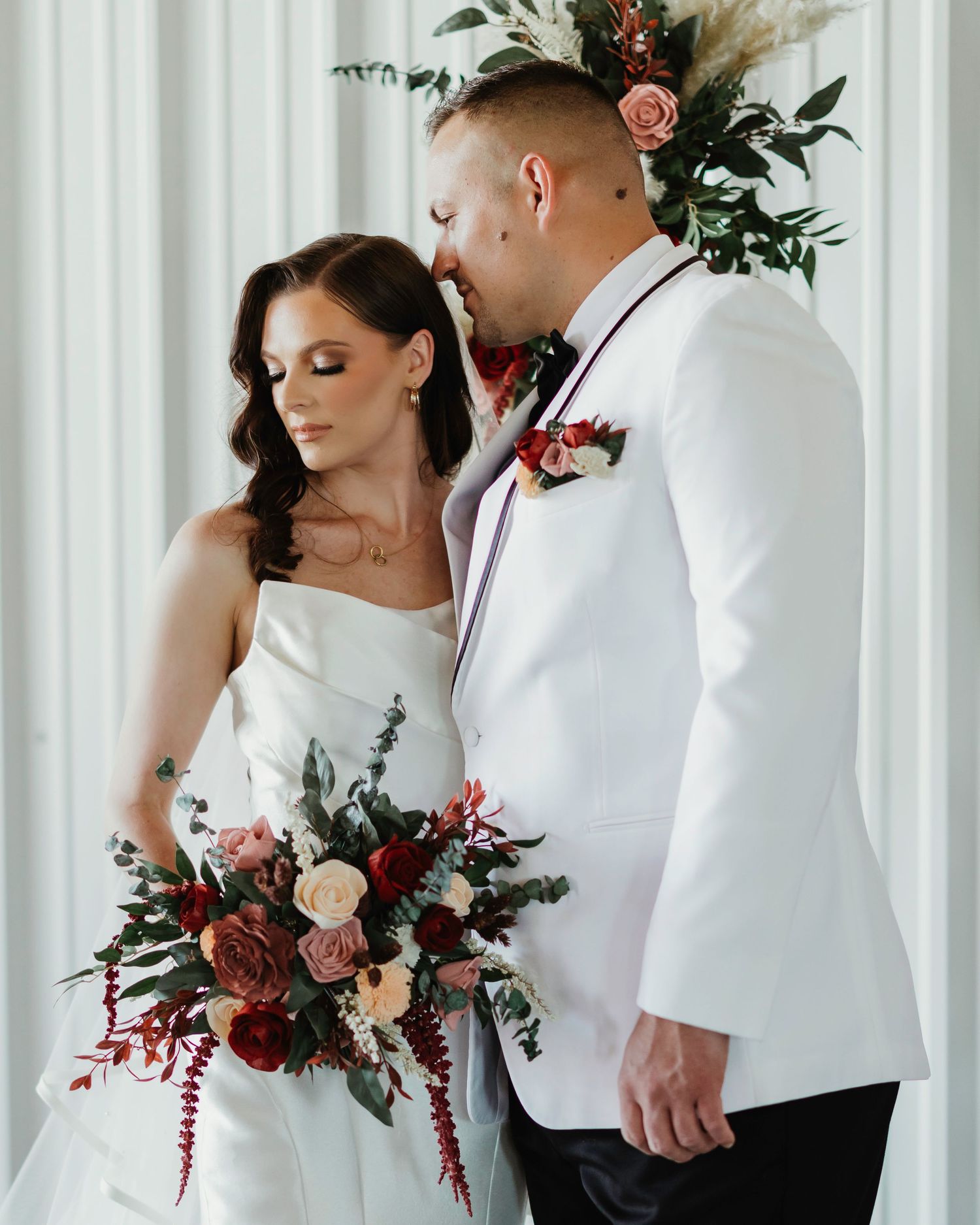 Elegant wedding couple embracing near floral arch with burgundy and blush arrangements.