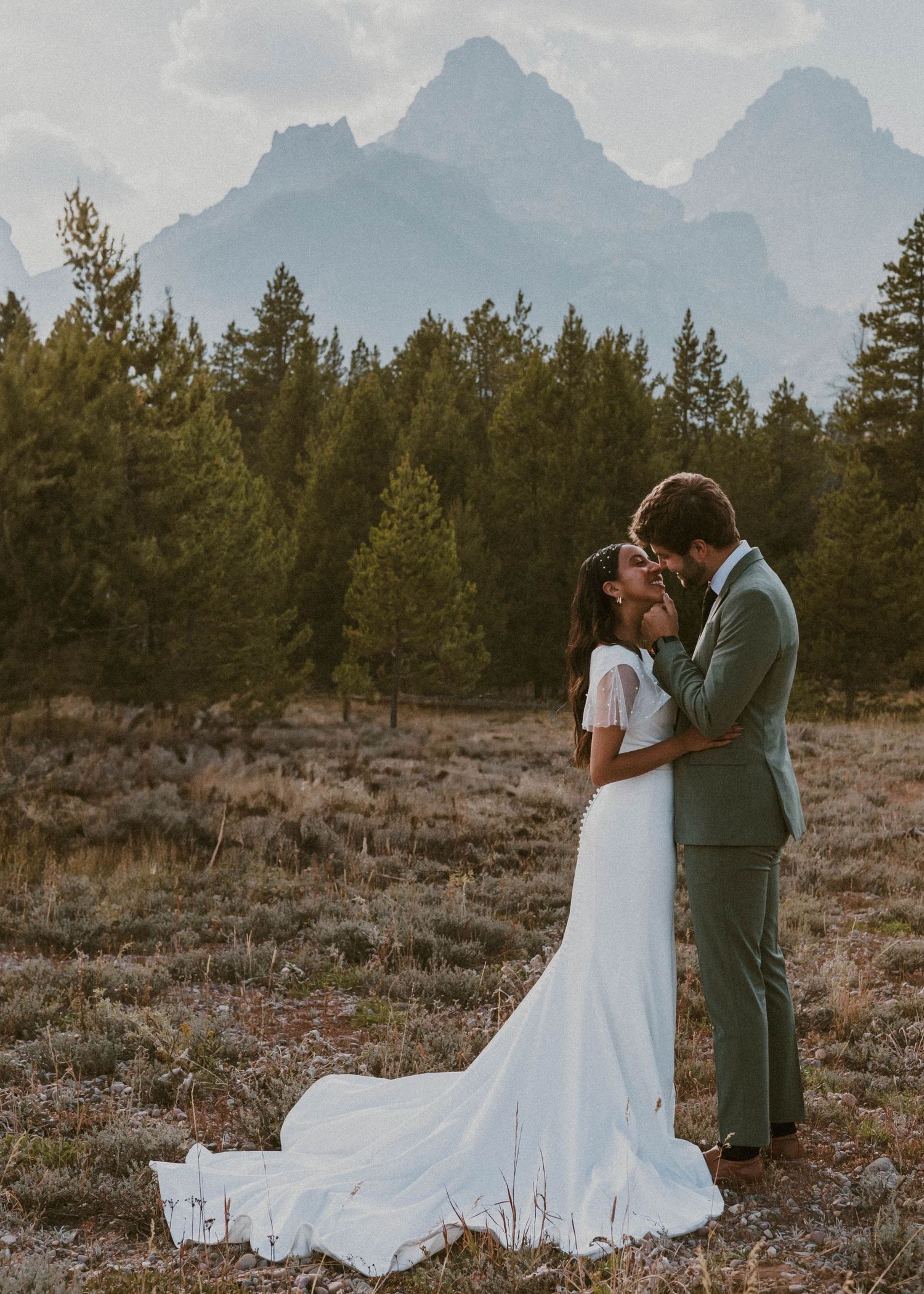 A tender moment between newlyweds against a backdrop of mountain peaks and pine trees at sunset.