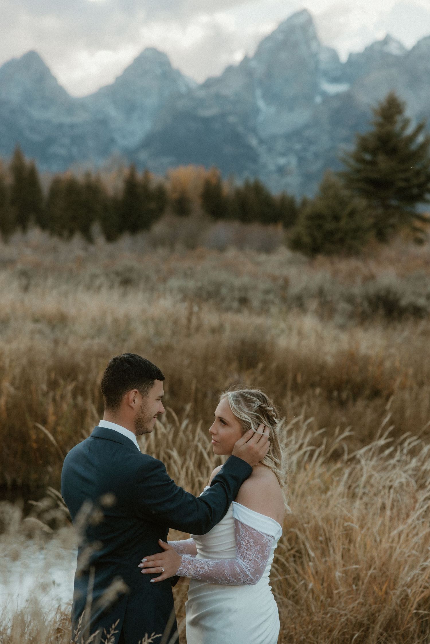 Newlyweds share an intimate moment in a field with dramatic mountain peaks in the background.