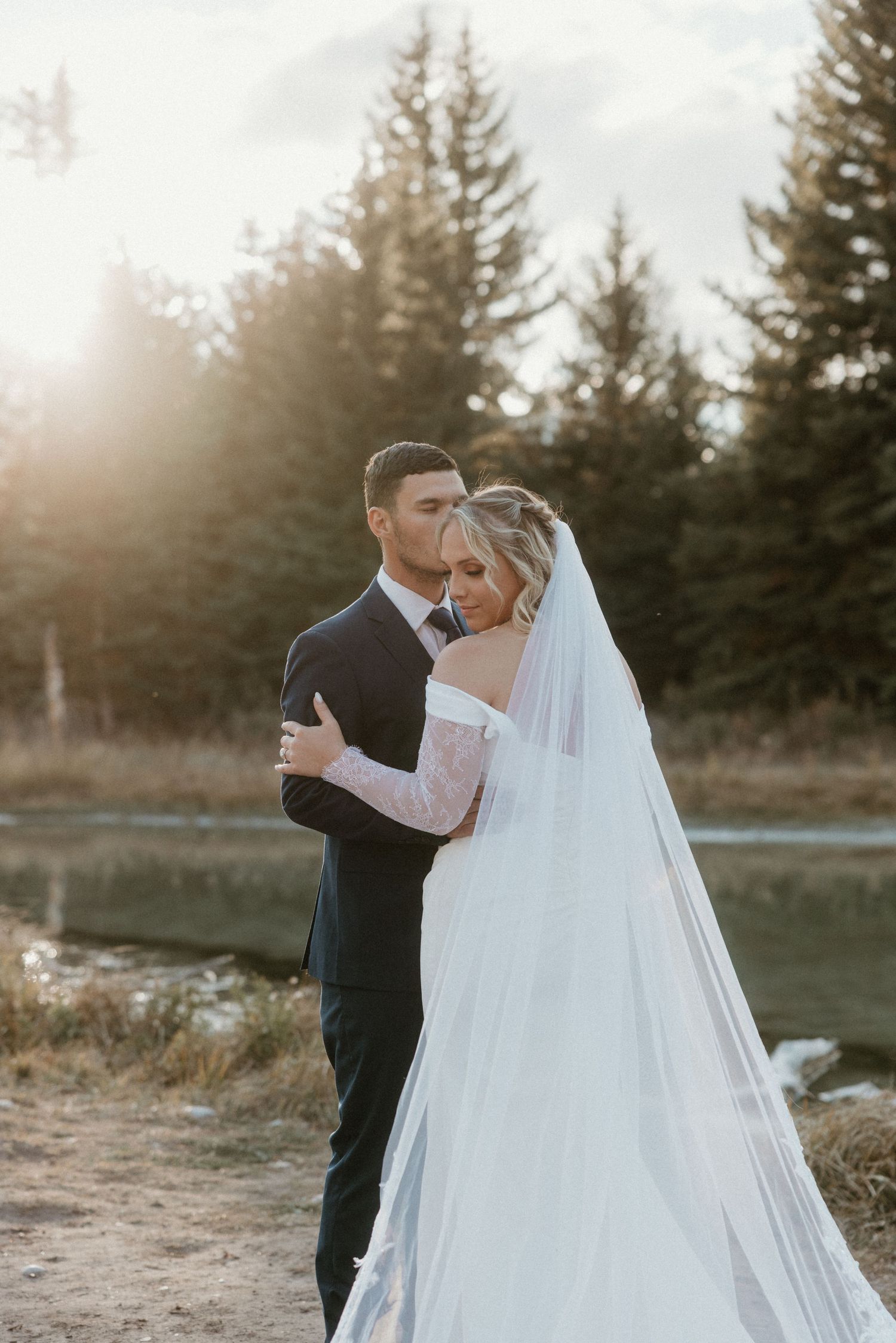 Newlyweds embrace by mountain lake with evergreen trees in background during sunset.