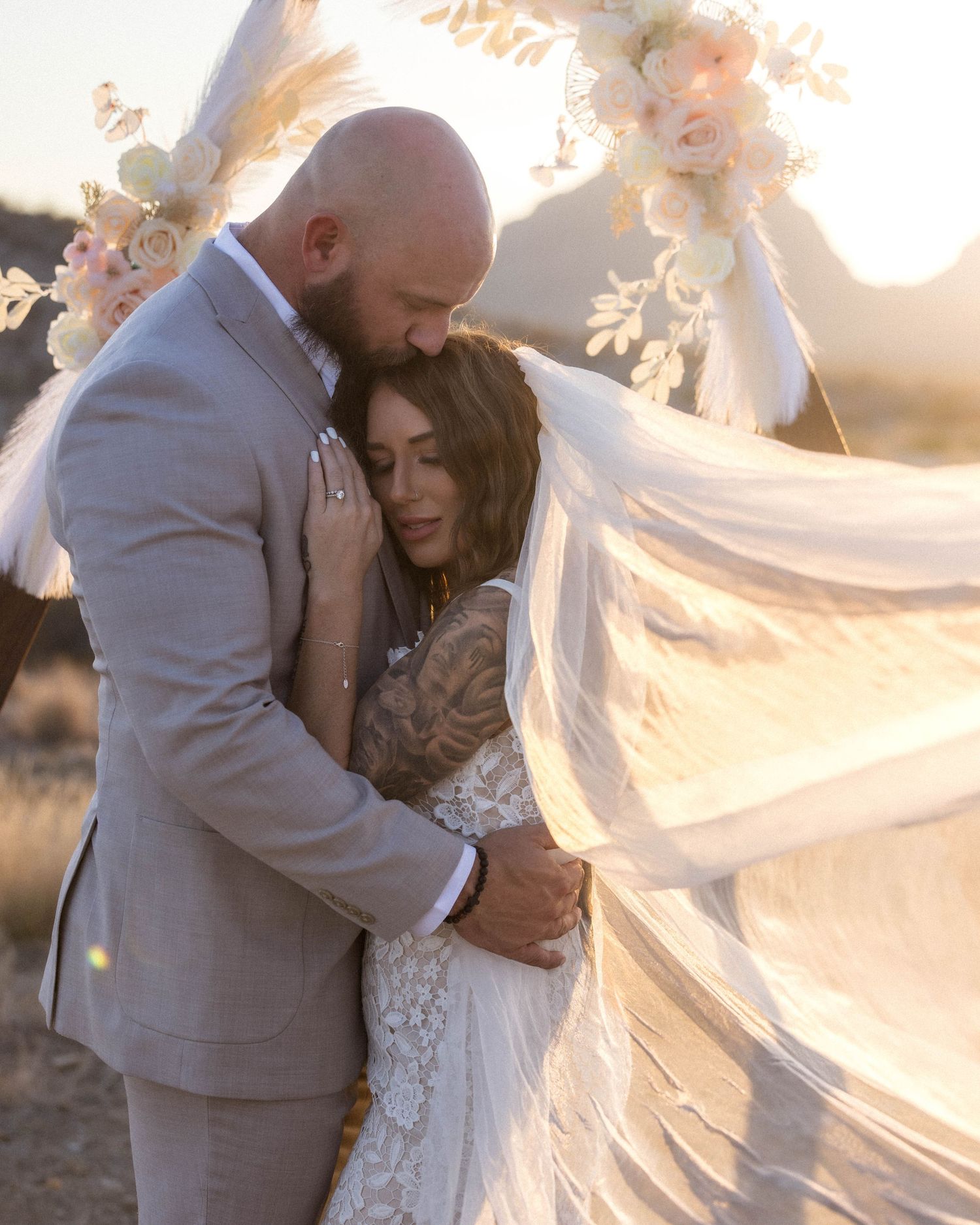 Romantic wedding couple embracing at sunset with flowing veil creating ethereal atmosphere in desert setting.