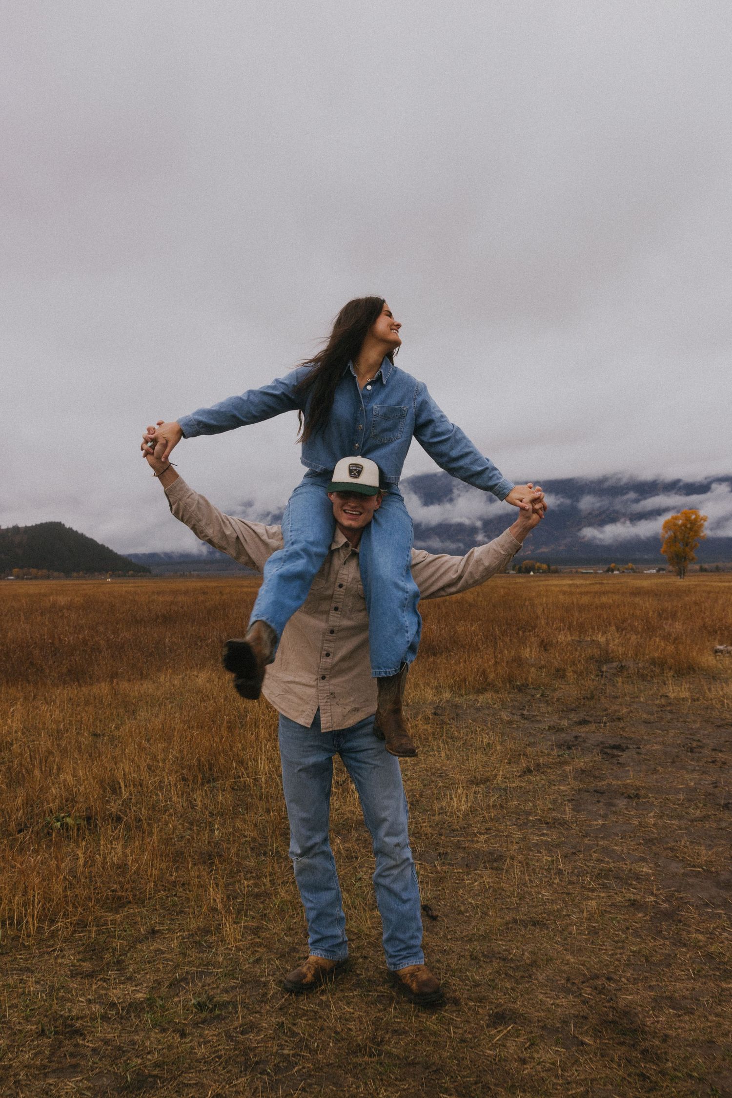 A couple performs a romantic dance lift in an open field with cloudy mountains behind them.