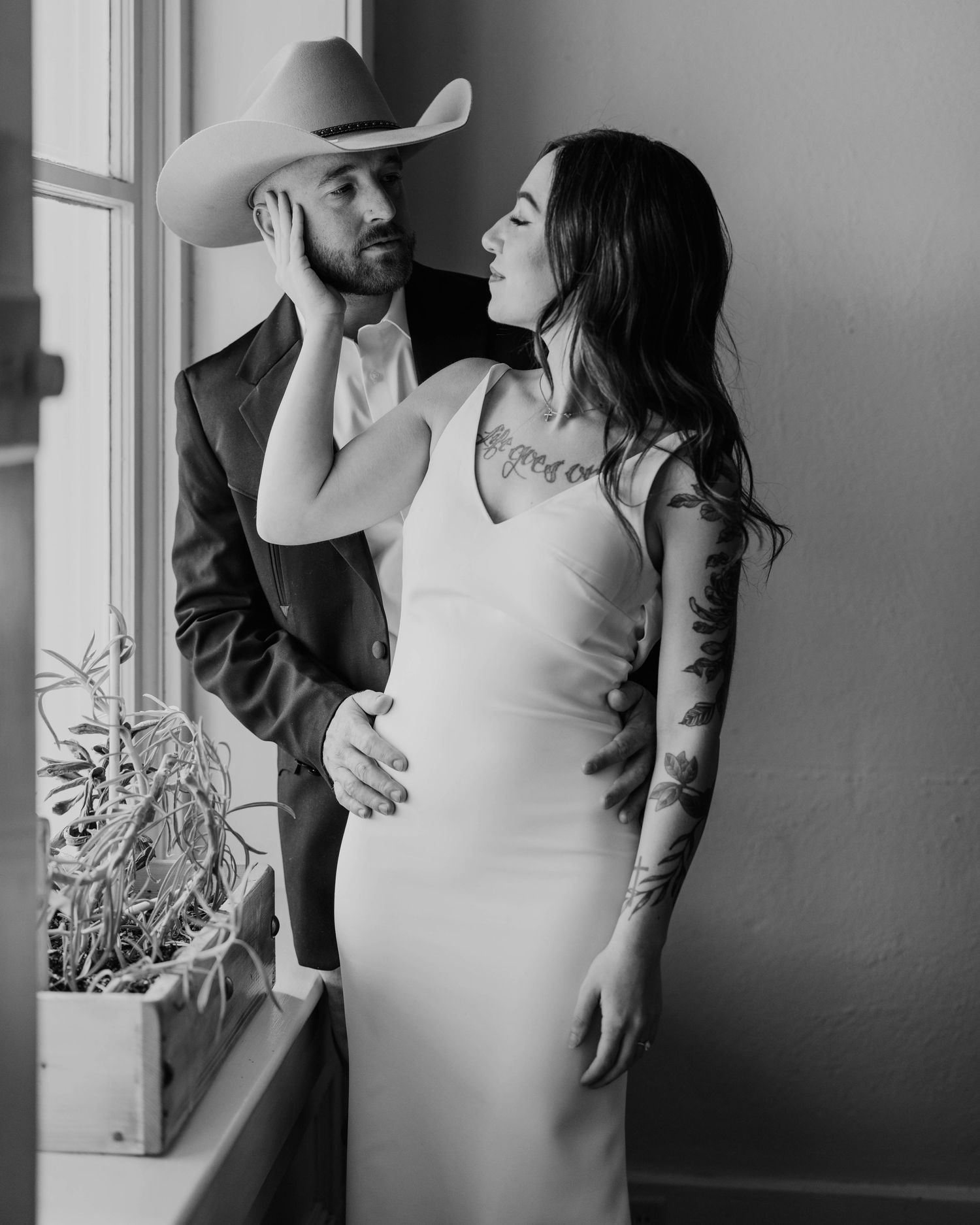 A romantic black and white portrait of a couple by a window, with one wearing a cowboy hat.