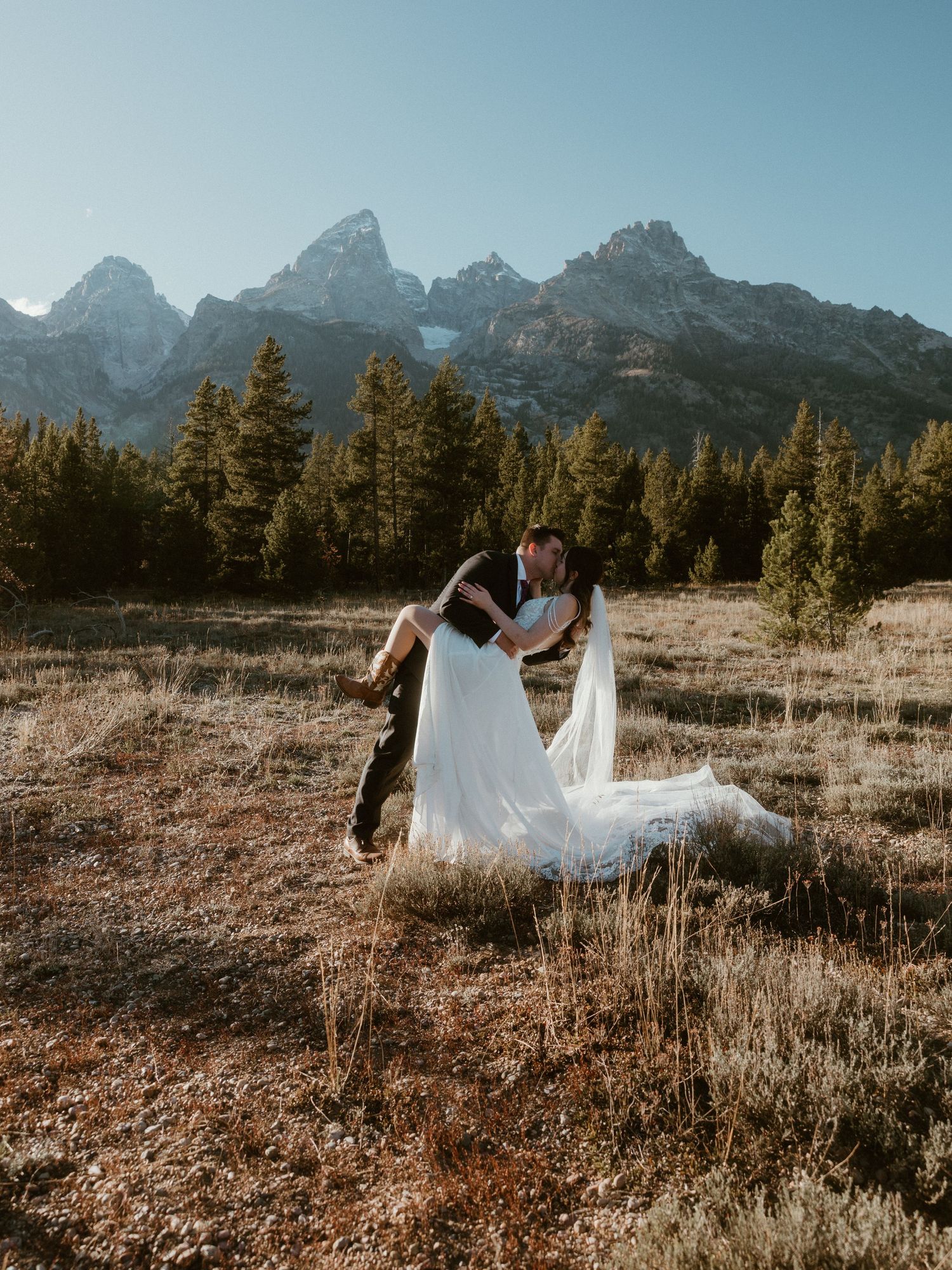 Bride and groom dancing playfully in mountain field during sunset.